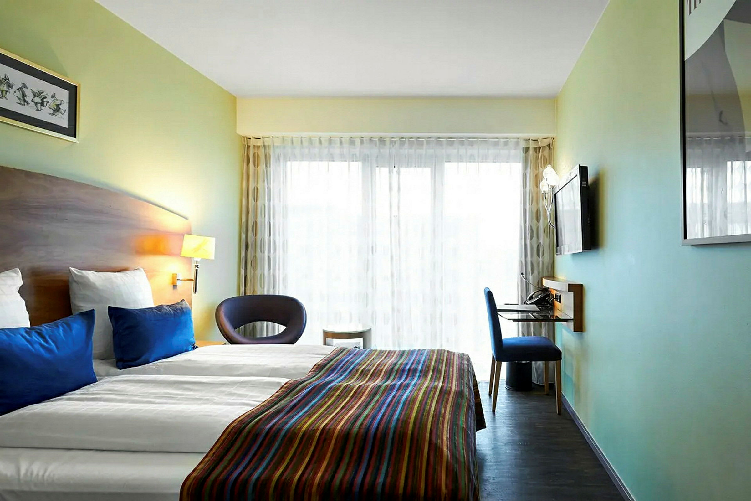 Ett modernt hotellrum med en dubbelsäng, blå kuddar och ett färgglatt överkast. Rummet har också en väggmonterad TV, ett skrivbord och en stol, samt stora fönster med ljusa gardiner. (AI-genererad text)