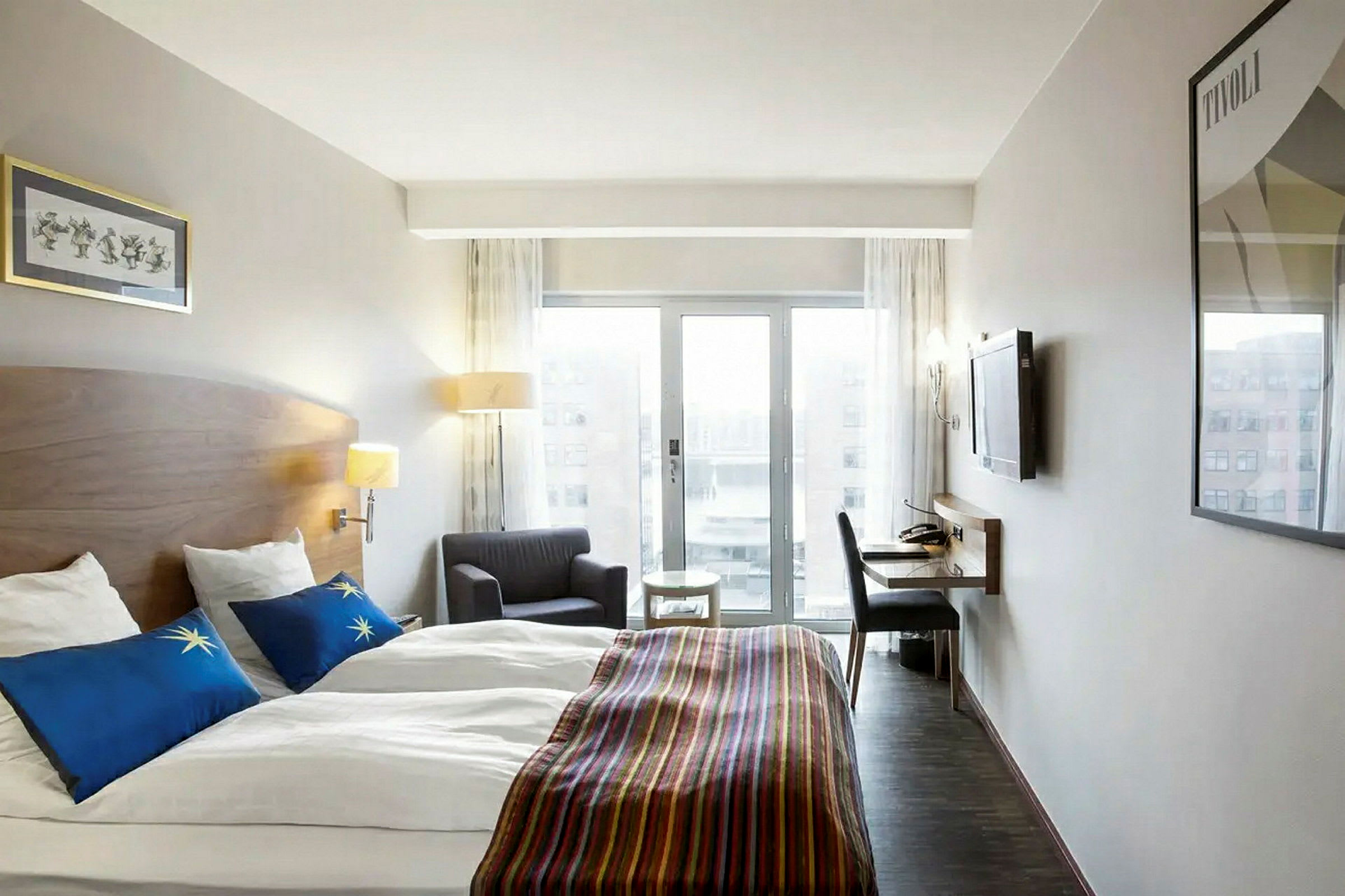Ett hotellrum med två enkelsängar, en fåtölj, ett skrivbord och en TV på väggen. Stora fönster släpper in dagsljus. (AI-genererad text)