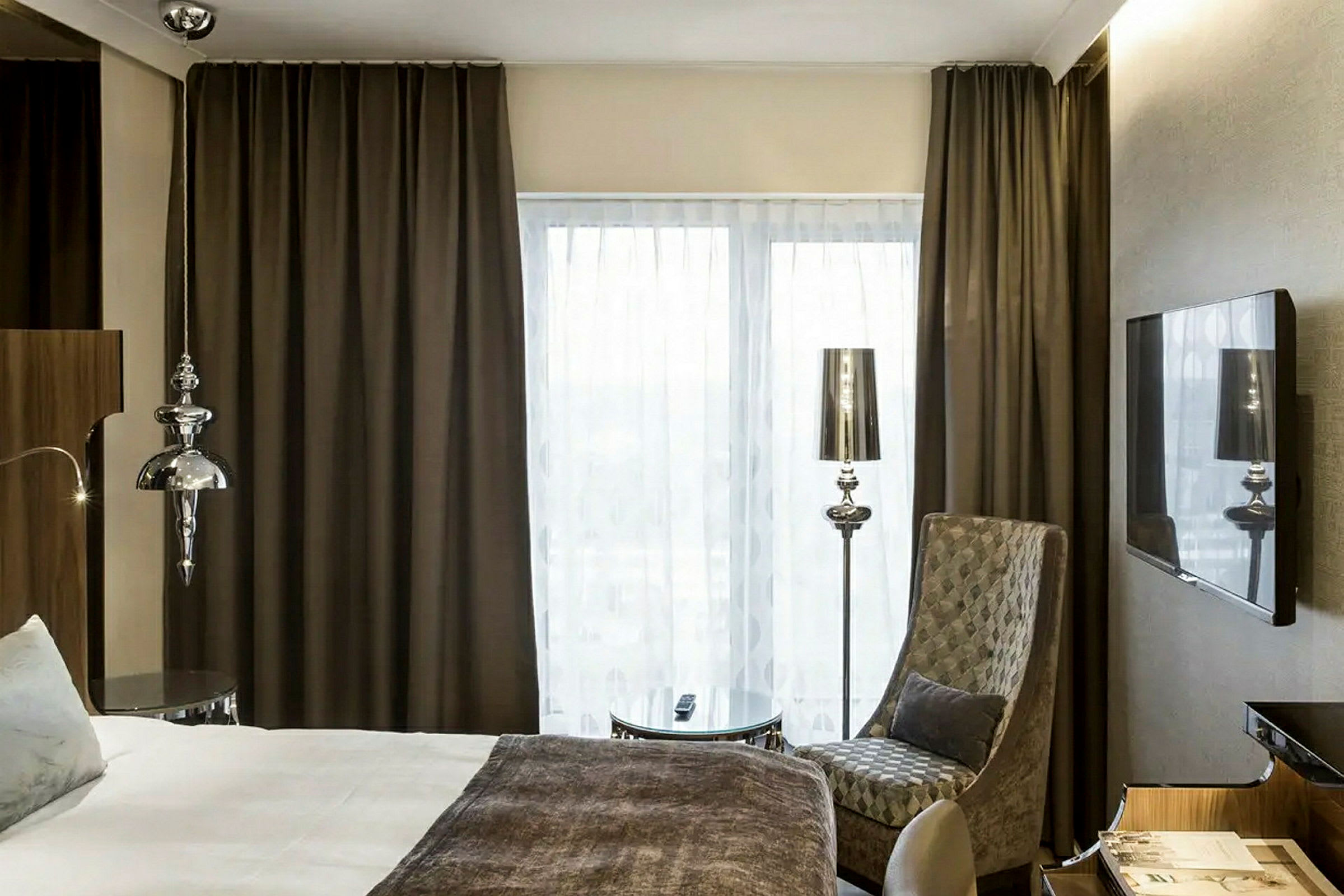 Ett hotellrum med en säng, en stol, ett soffbord, gardiner och en TV på väggen. (AI-genererad text)