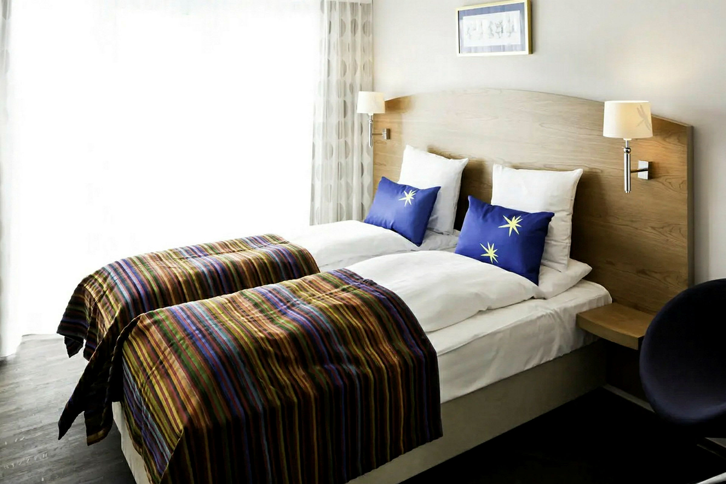 Hotellrum med två enkelsängar som har träpanelad sänggavel, randiga överkast och blå kuddar med stjärnmotiv. (AI-genererad text)