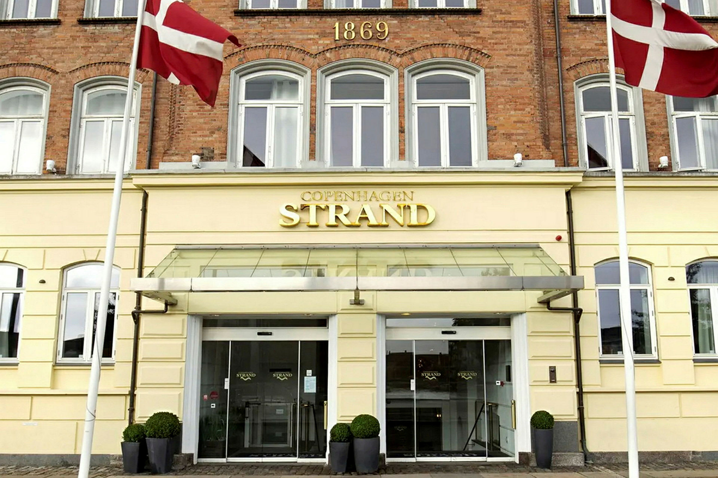 En byggnad med gul fasad och en skylt där det står "Copenhagen Strand" ovanför entrédörrarna. Två danska flaggor hänger på vardera sida. (AI-genererad text)