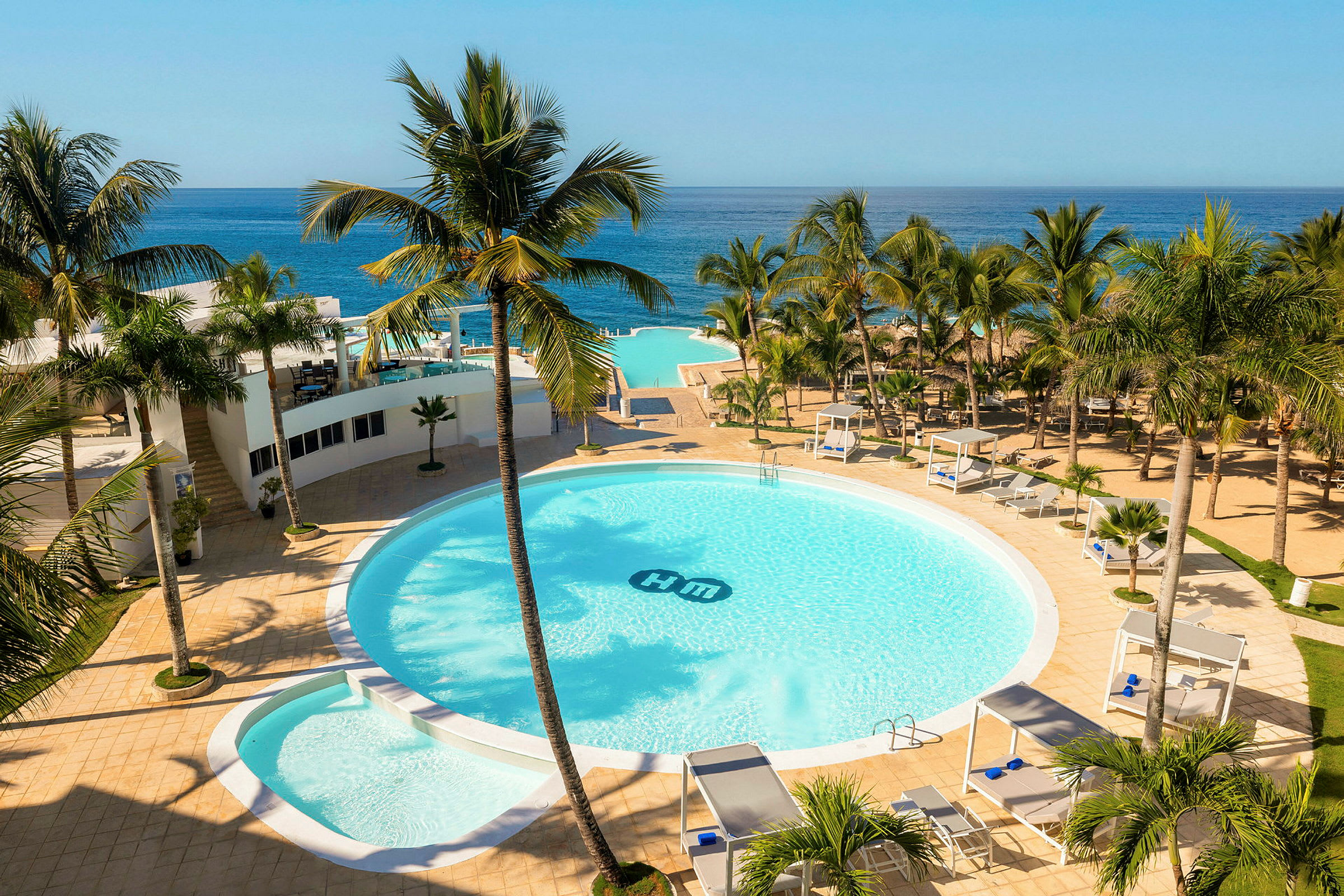 Ett hotellområde med en stor rund pool omgiven av palmer, solsängar och cabanas. Utsikt över havet i bakgrunden. (AI-genererad text)