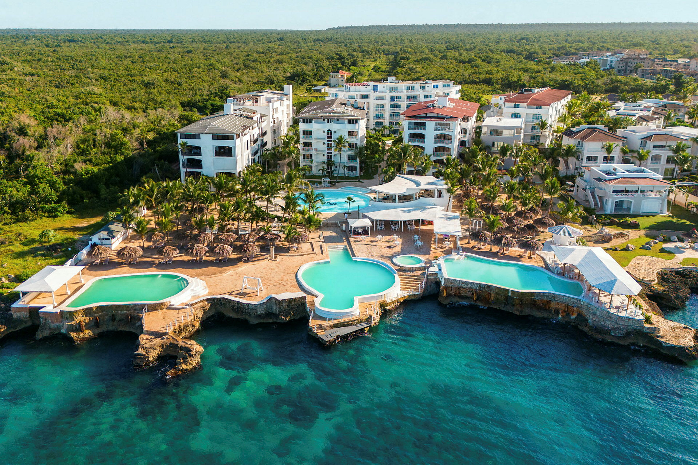 En strandnära semesterort med flera vita byggnader, pooler och palmer, belägen intill en klippa vid blått hav. (AI-genererad text)