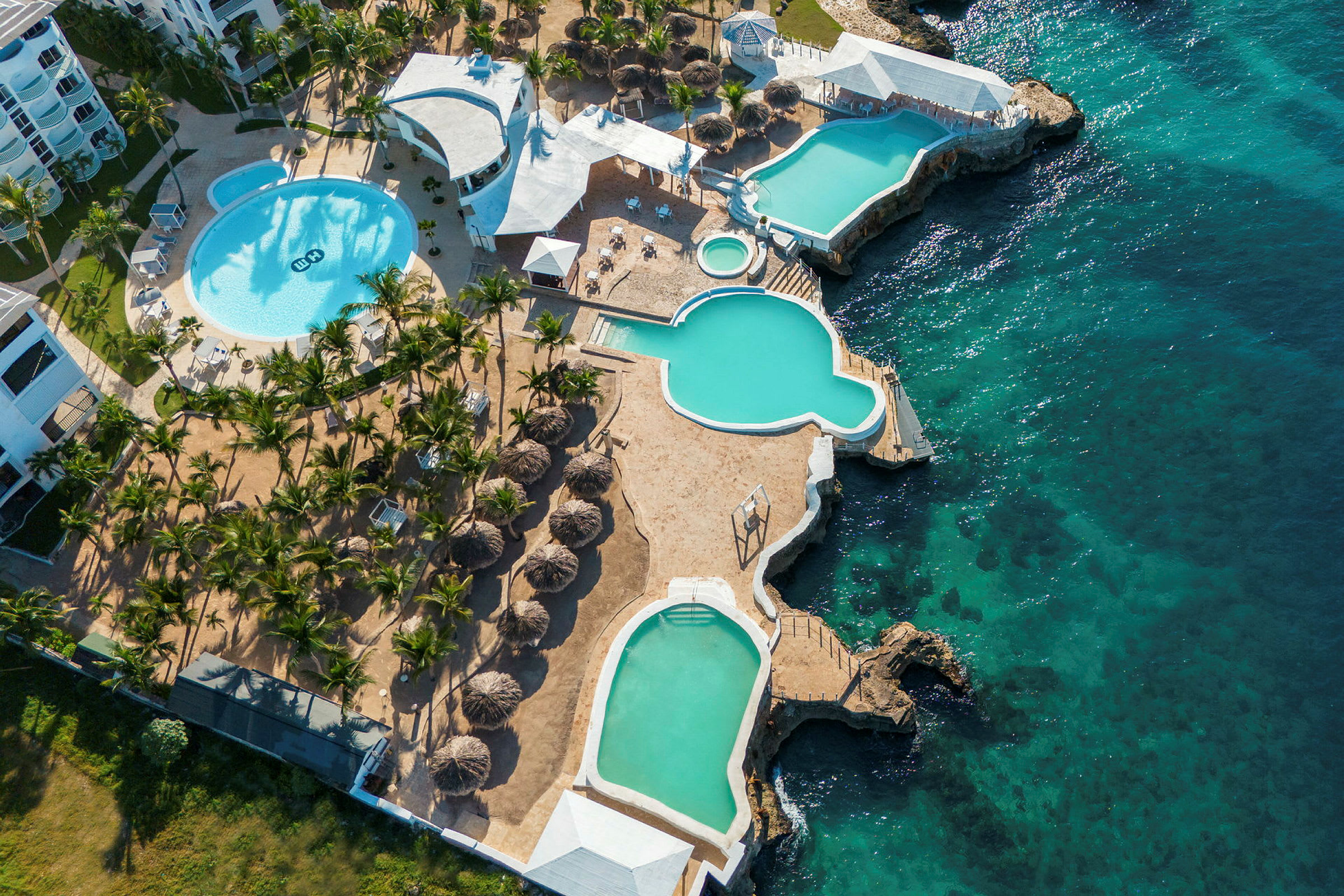Flygbild över en resort med flera pooler, omgivna av solstolar och palmer, intill ett klart blått hav. (AI-genererad text)