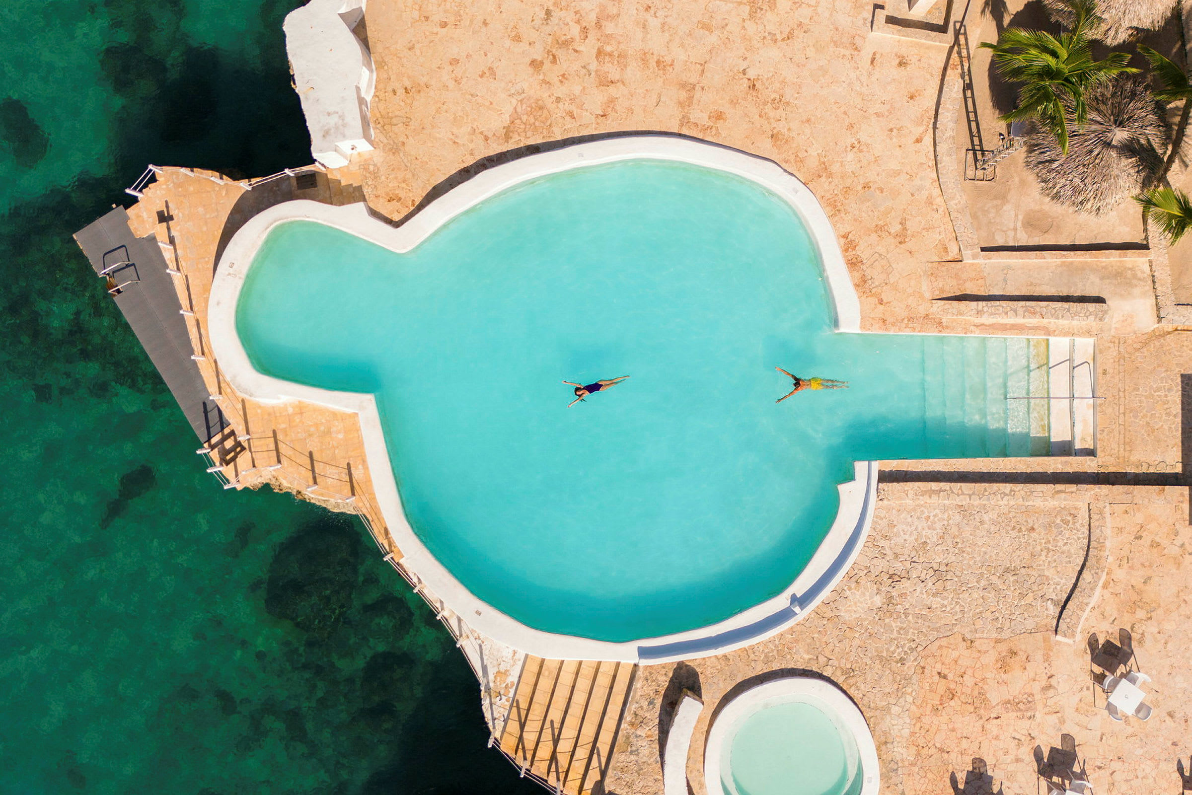 Vy ovanifrån av en pool med två personer som flyter på rygg. Poolområdet är omringat av stenplattor vid en kust. (AI-genererad text)