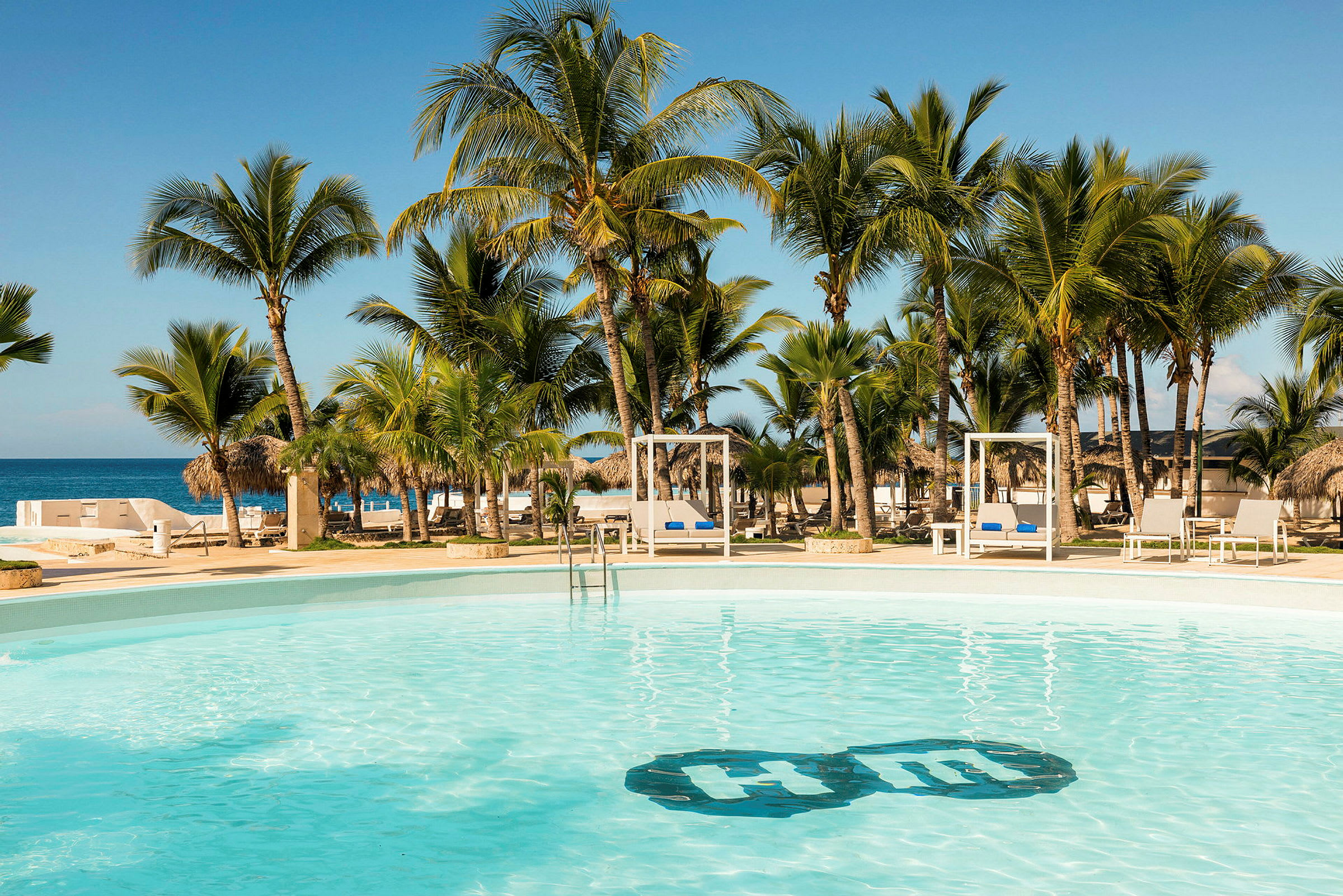 En pool omgiven av palmer med utsikt över stranden och havet under en klarblå himmel. (AI-genererad text)