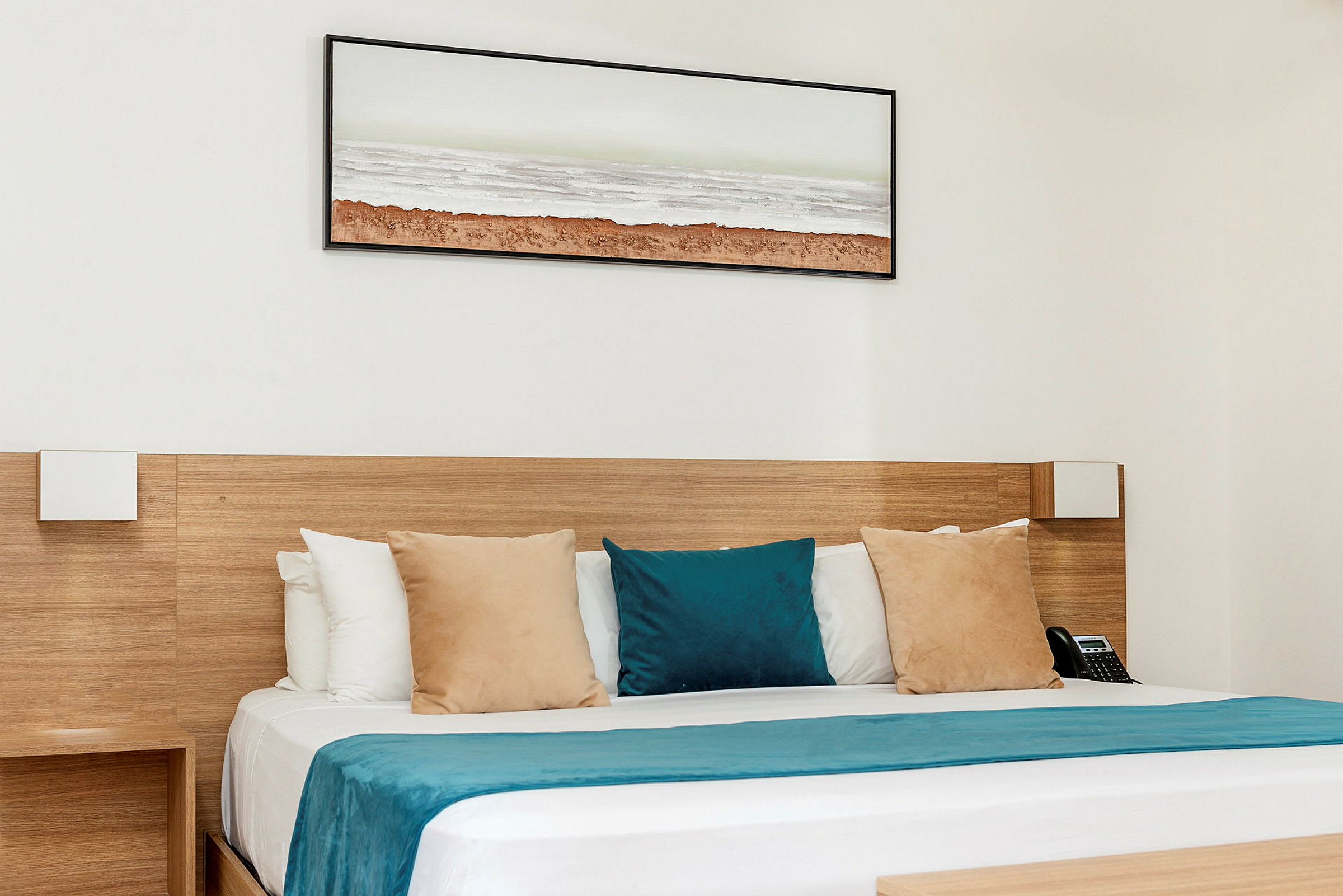 Ett hotellrum med en säng som har kuddar i beige och blå färger och en blå sängöverkast. En tavla med ett landskapsmotiv är placerad på väggen ovanför. (AI-genererad text)