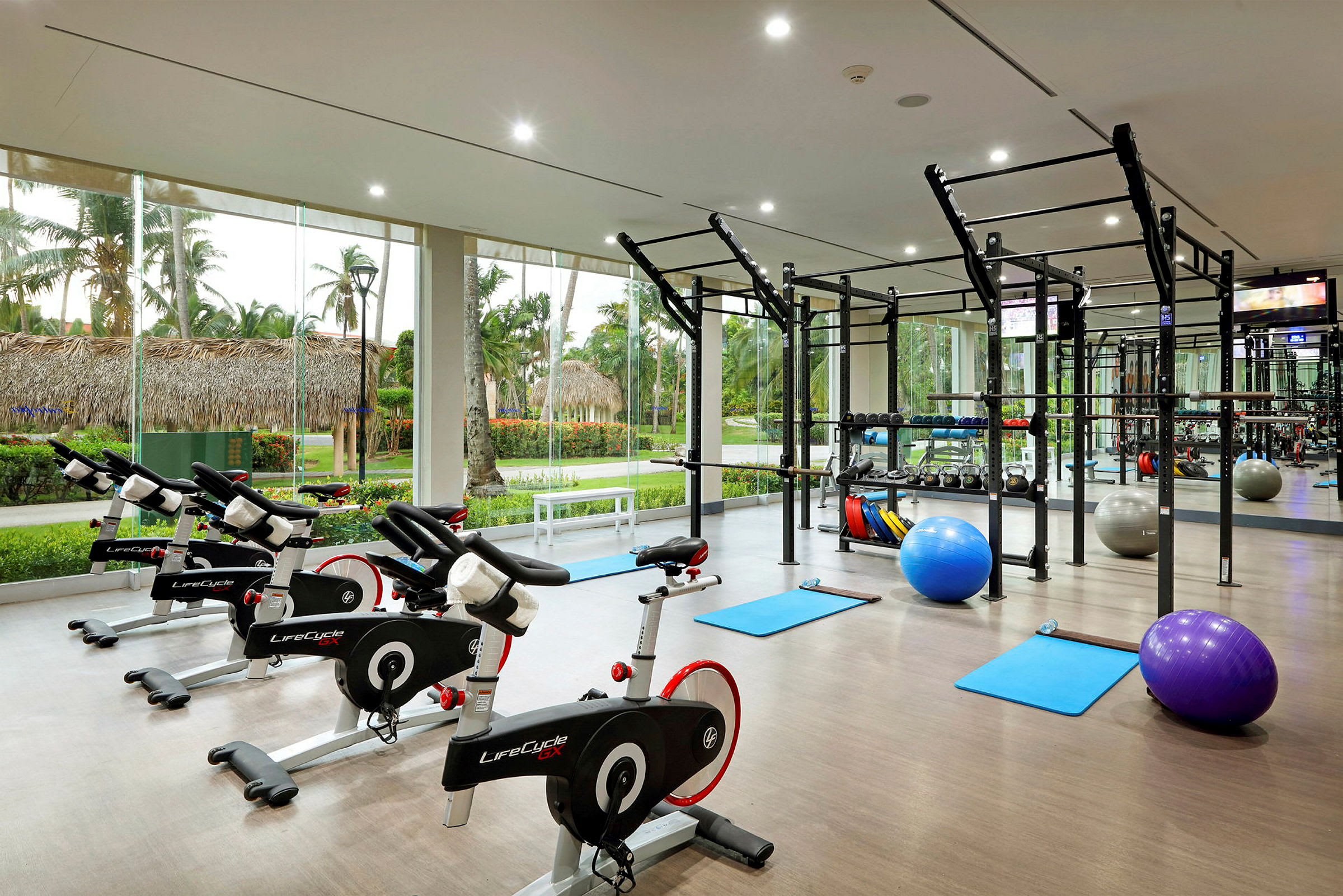 Ett modernt gym med träningscyklar, yogamattor, träningsbollar och styrketräningsutrustning, med stora fönster som ger utsikt över en tropisk trädgård. (AI-genererad text)