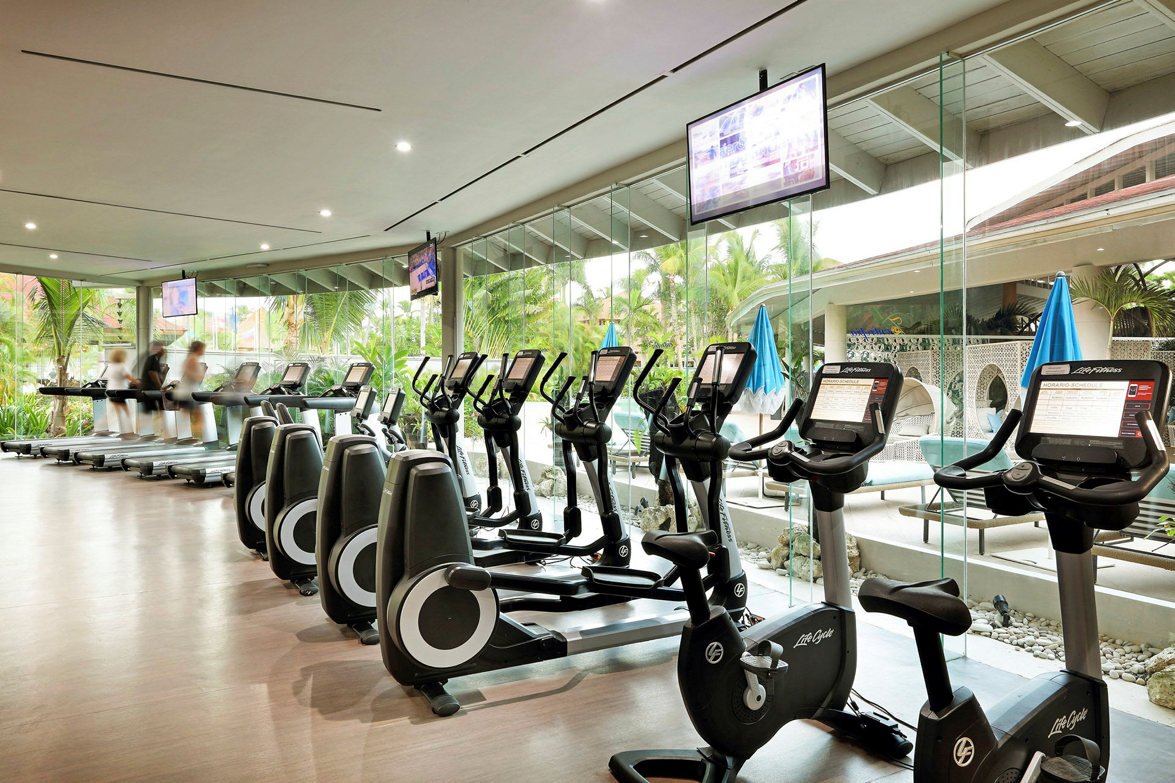 Ett gym med träningsmaskiner inklusive löpband och motionscyklar. Stora fönster visar grönska och en utomhusmiljö. (AI-genererad text)