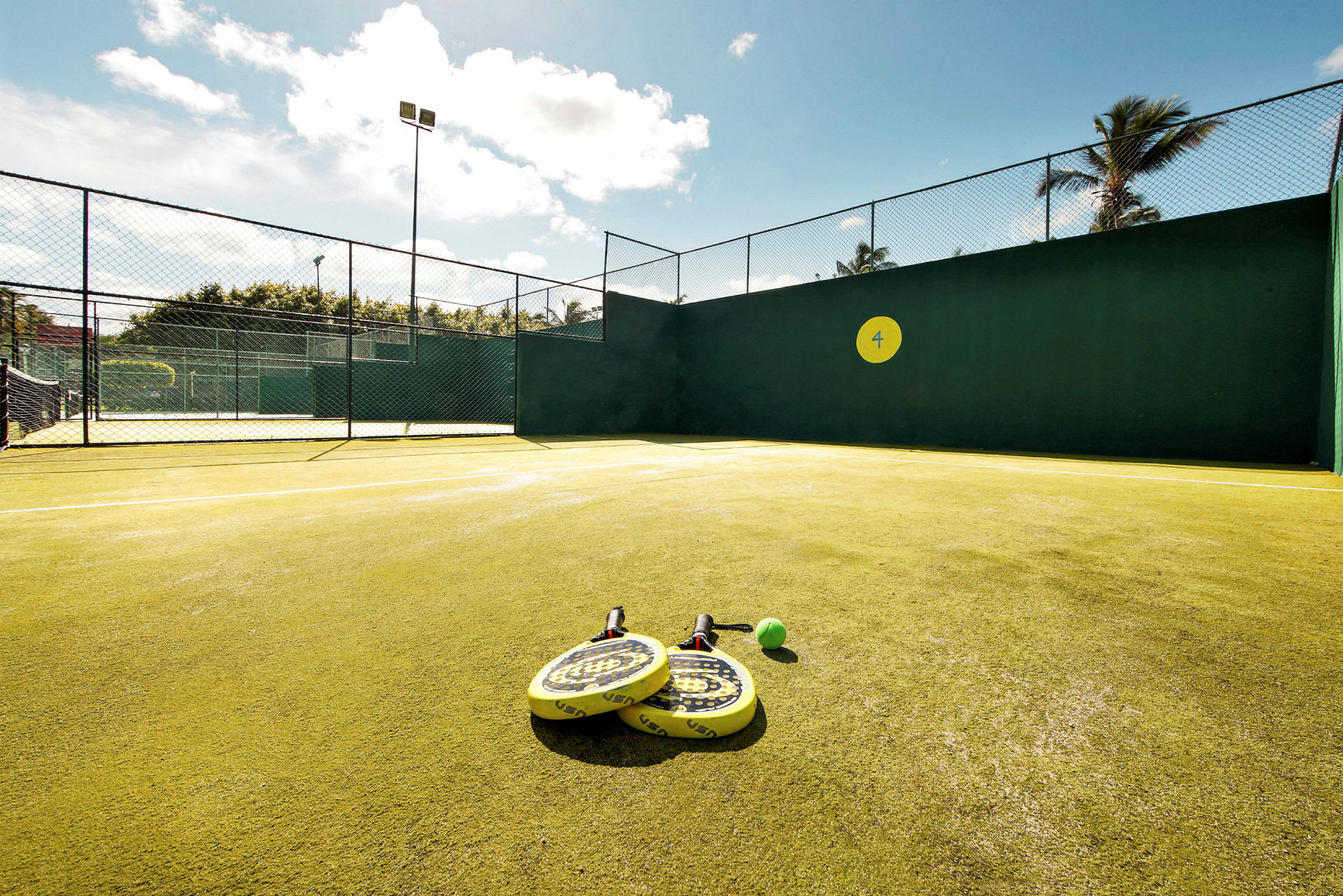 En padelbana med gula racketar och en boll placerade på marken. Soligt väder. (AI-genererad text)