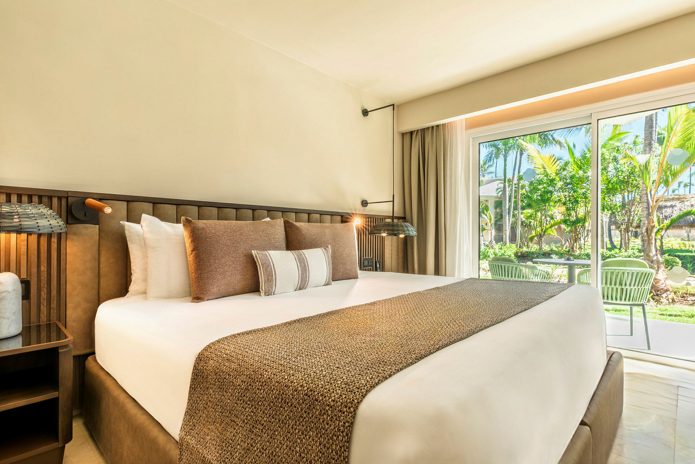 Ett hotellrum med en stor säng utrustad med flera kuddar, sängbord och en glasad dörr som leder till en uteplats och gröna omgivningar. (AI-genererad text)