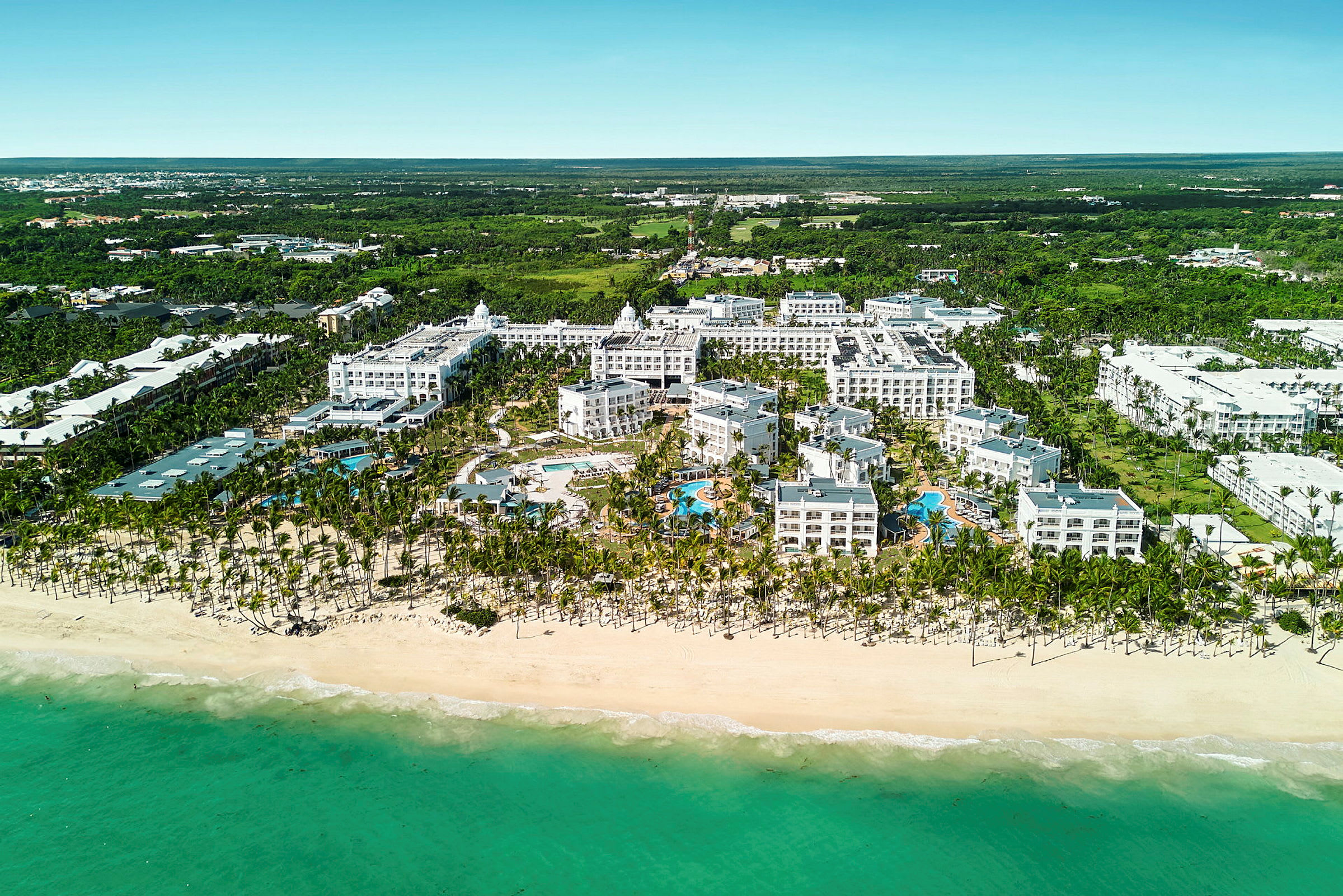 Flygbild av ett hotellkomplex vid en strand inklusive poolområde, omgiven av grönska. (AI-genererad text)