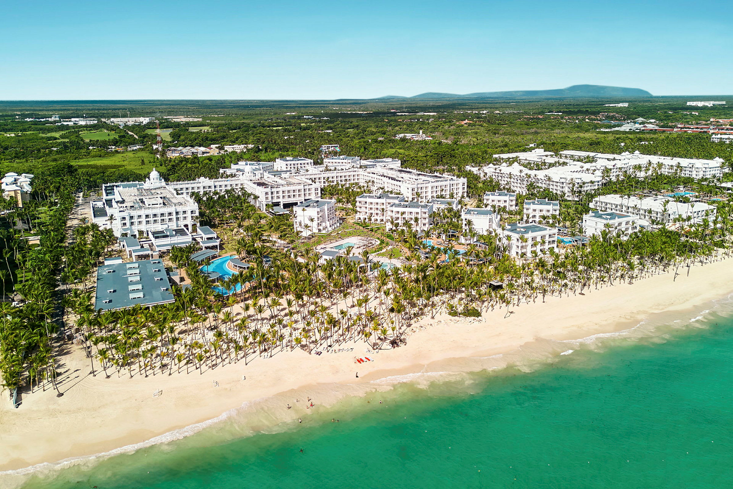 Flygvy över hotellkomplex vid en sandstrand med palmer och turkost hav. (AI-genererad text)