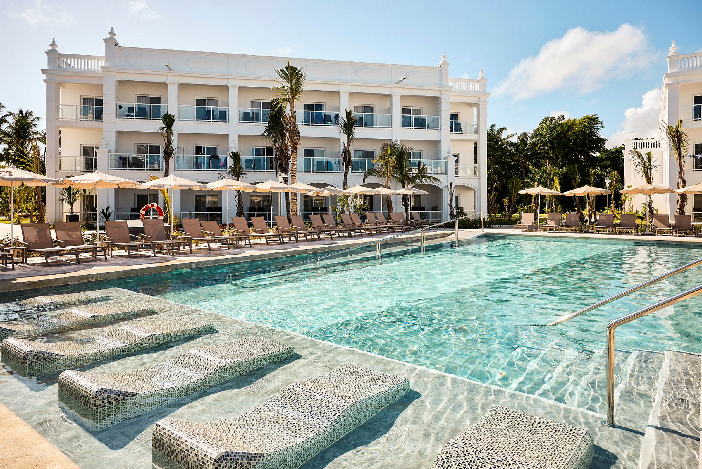 En swimmingpool framför ett hotell med vit fasad och flera våningar. (AI-genererad text)