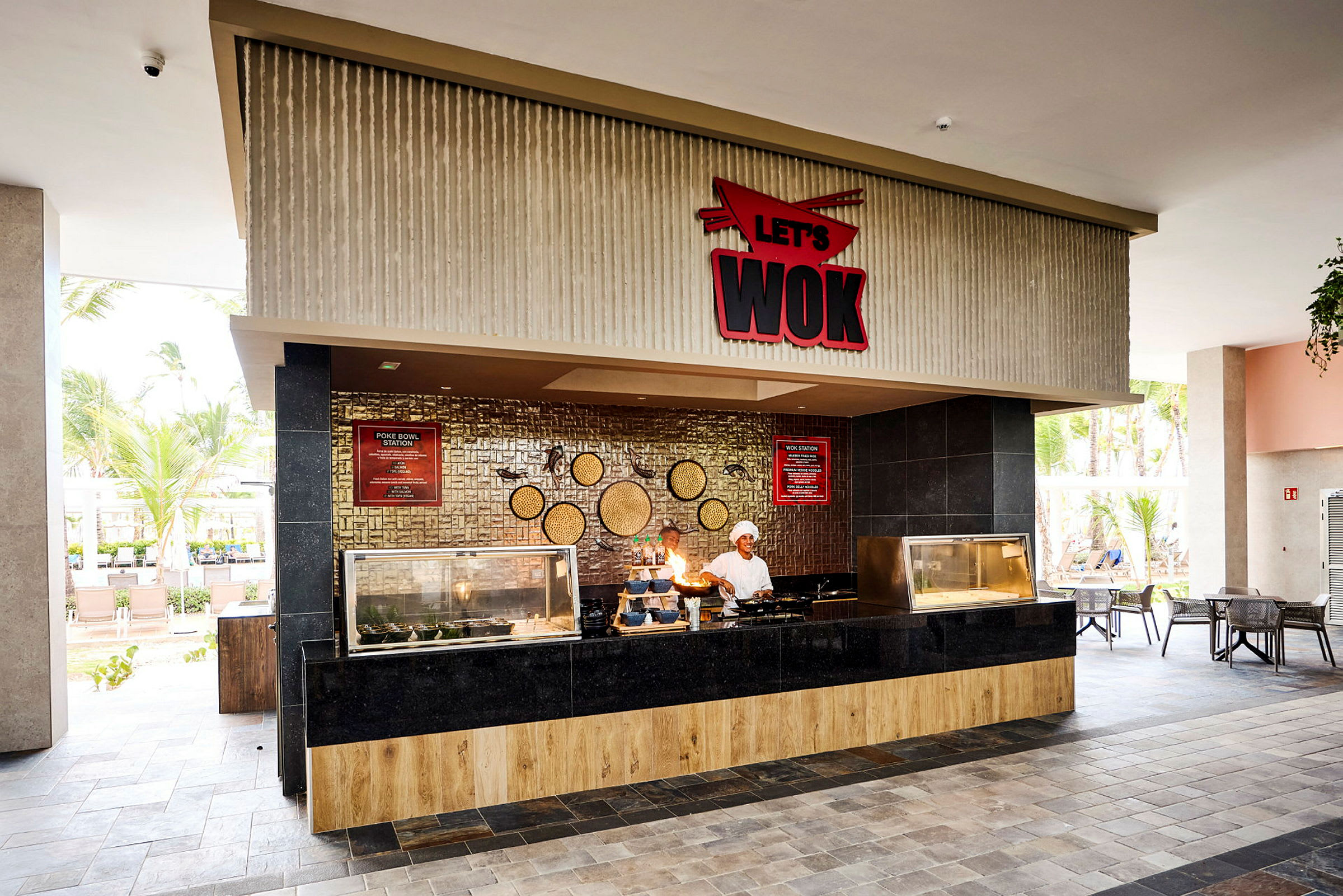 Bild av en restaurang med en serveringsdisk märkt 'Let's Wok'. Interiören har en modern design med trä och svart panel, och det finns en utomhusmiljö med palmer i bakgrunden. (AI-genererad text)
