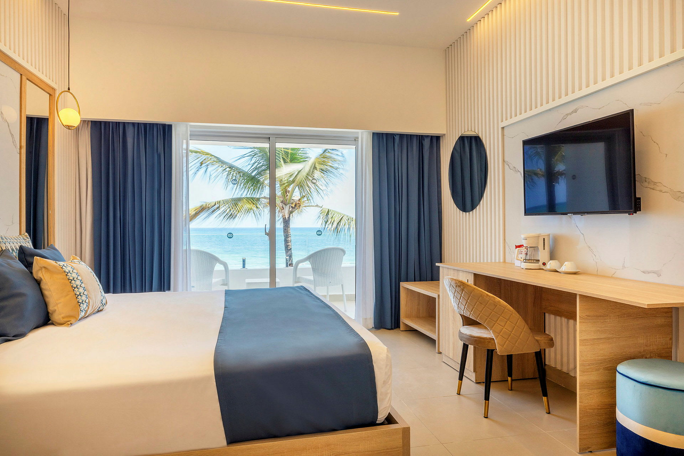 Ett hotellrum med en dubbelsäng, fönster med utsikt mot havet och en palm, samt inredning i ljusa färger. (AI-genererad text)