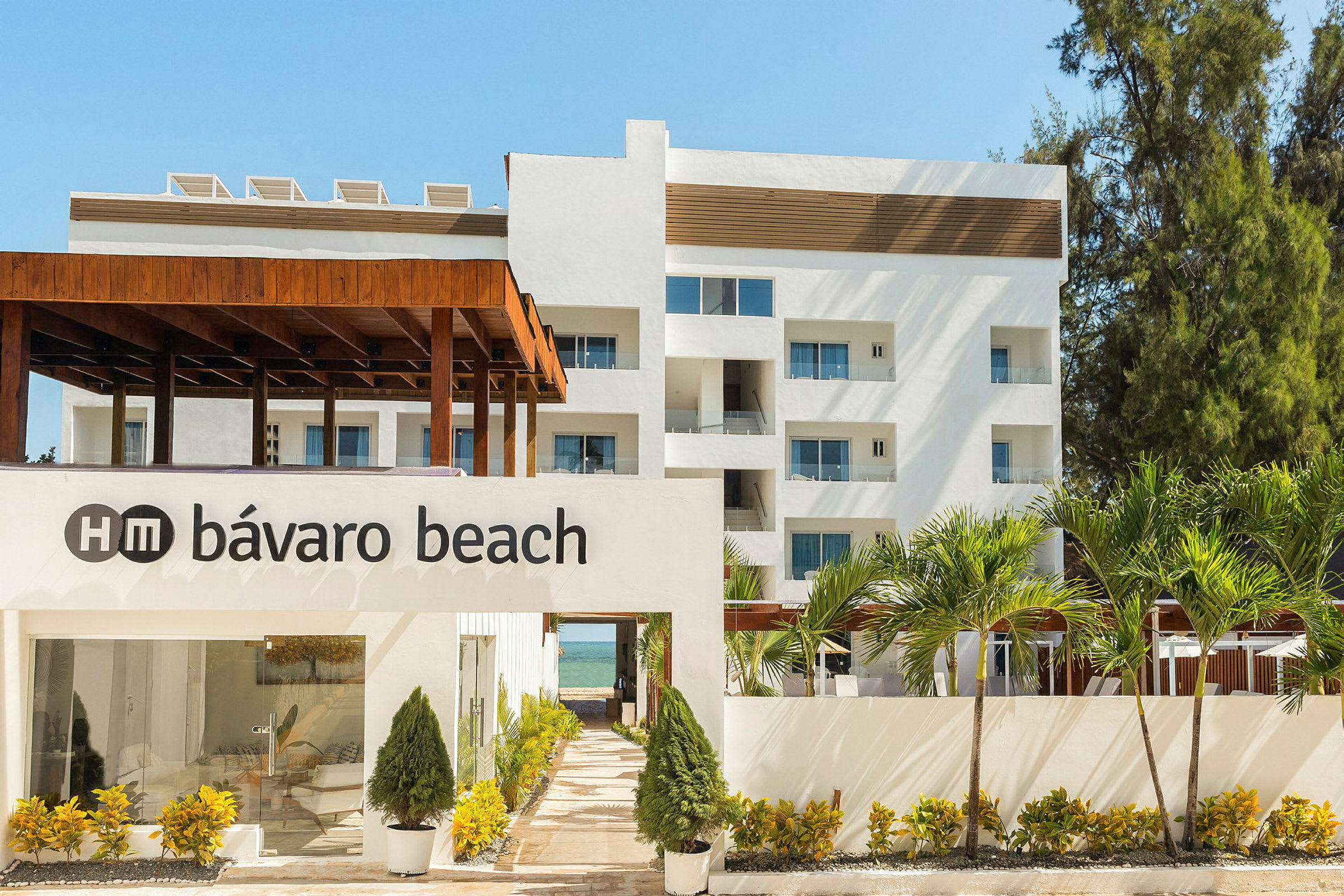 Bild av en vit byggnad med texten "HM Bávaro Beach" på en vägg framför, omgiven av frodig grönska och palmer. (AI-genererad text)