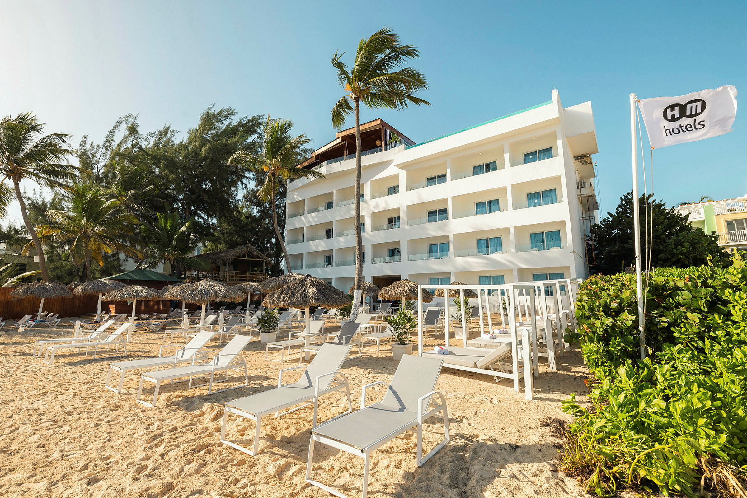En vit hotellbyggnad vid en sandstrand omgiven av parasoller och solstolar. (AI-genererad text)