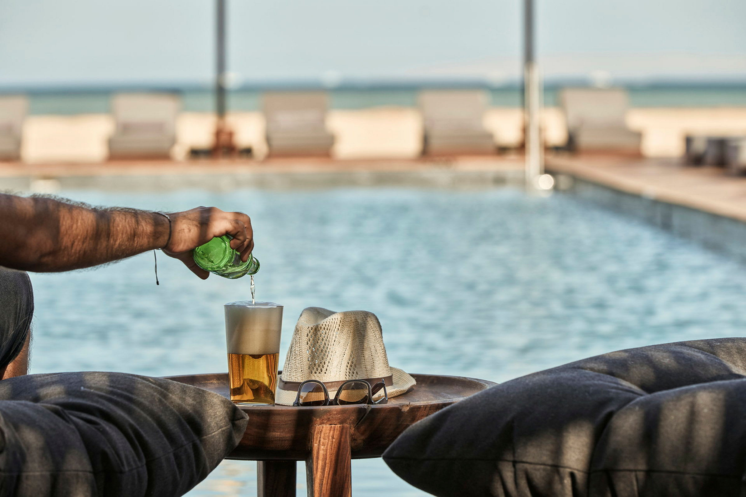 En person häller dryck från en grön flaska i ett glas vid poolen, med en hatt och glasögon på ett bord. (AI-genererad text)