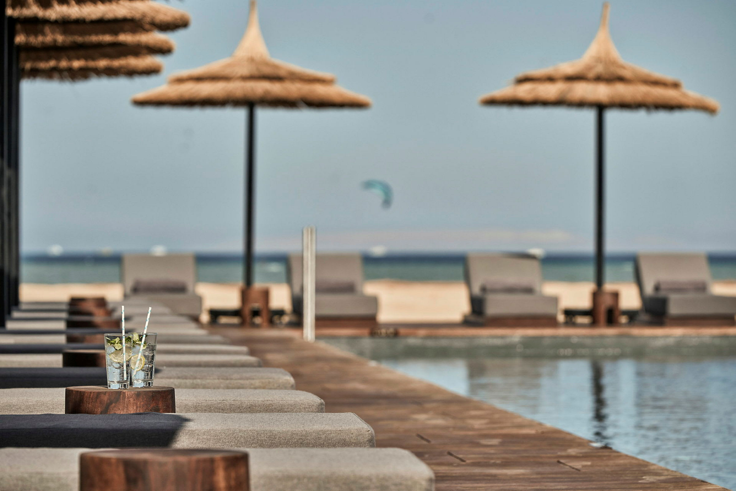 Solsängar med parasoller vid en pool, med stranden och havet i bakgrunden. (AI-genererad text)