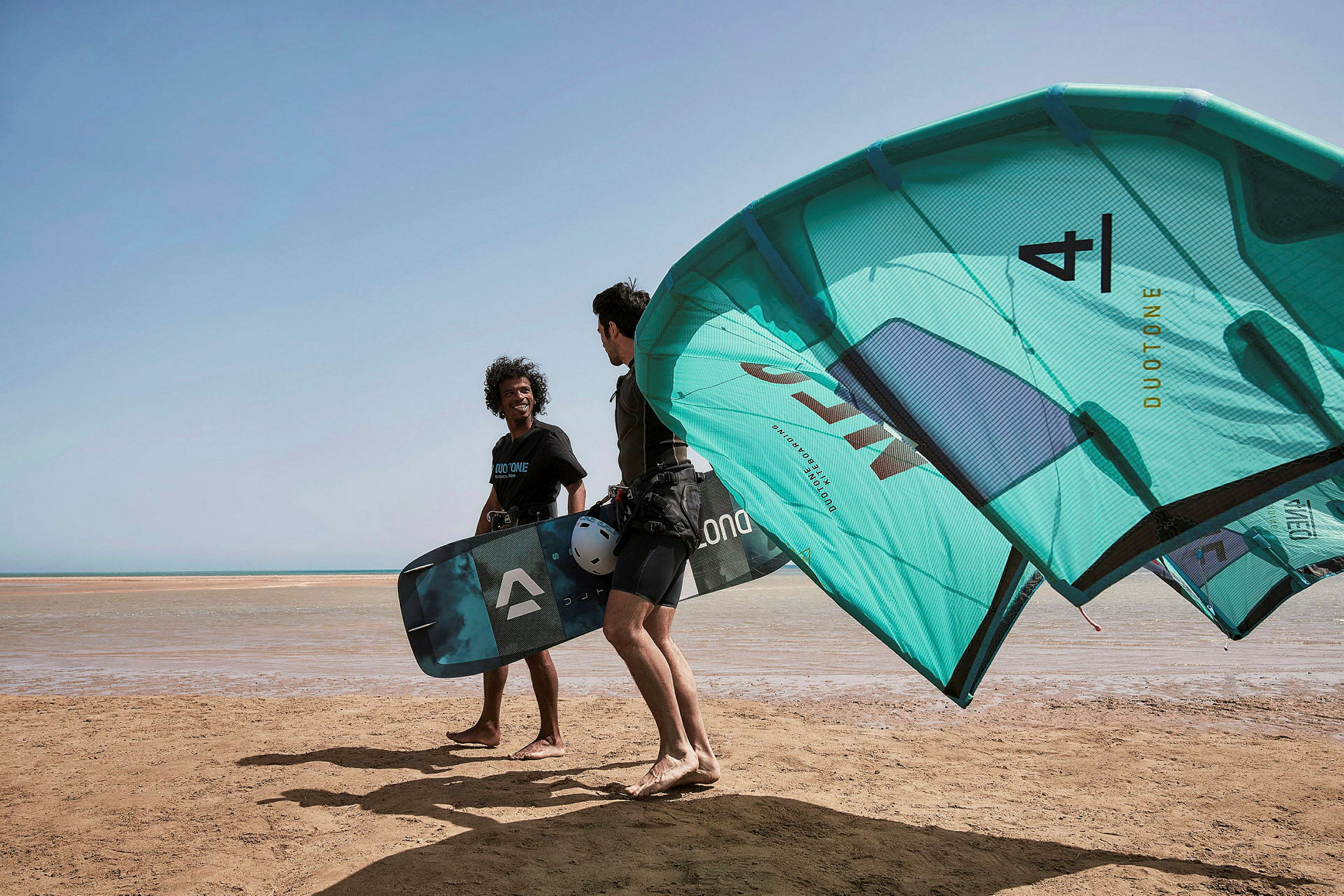 Två personer med kiteutrustning står på en strand. (AI-genererad text)