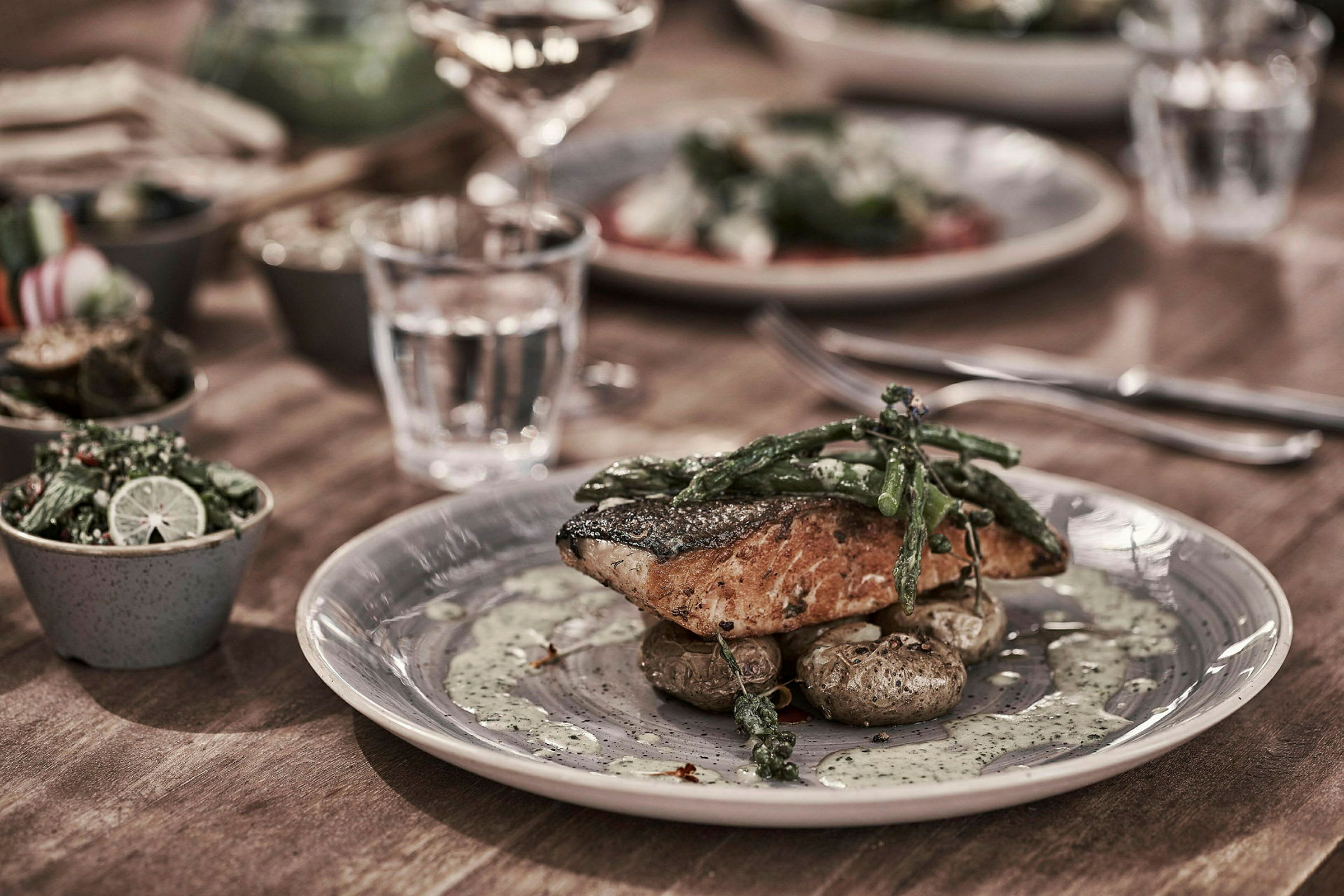 En tallrik med tillagad lax, potatis och grönsaker. I bakgrunden syns fler tillbehör och en dukning. (AI-genererad text)