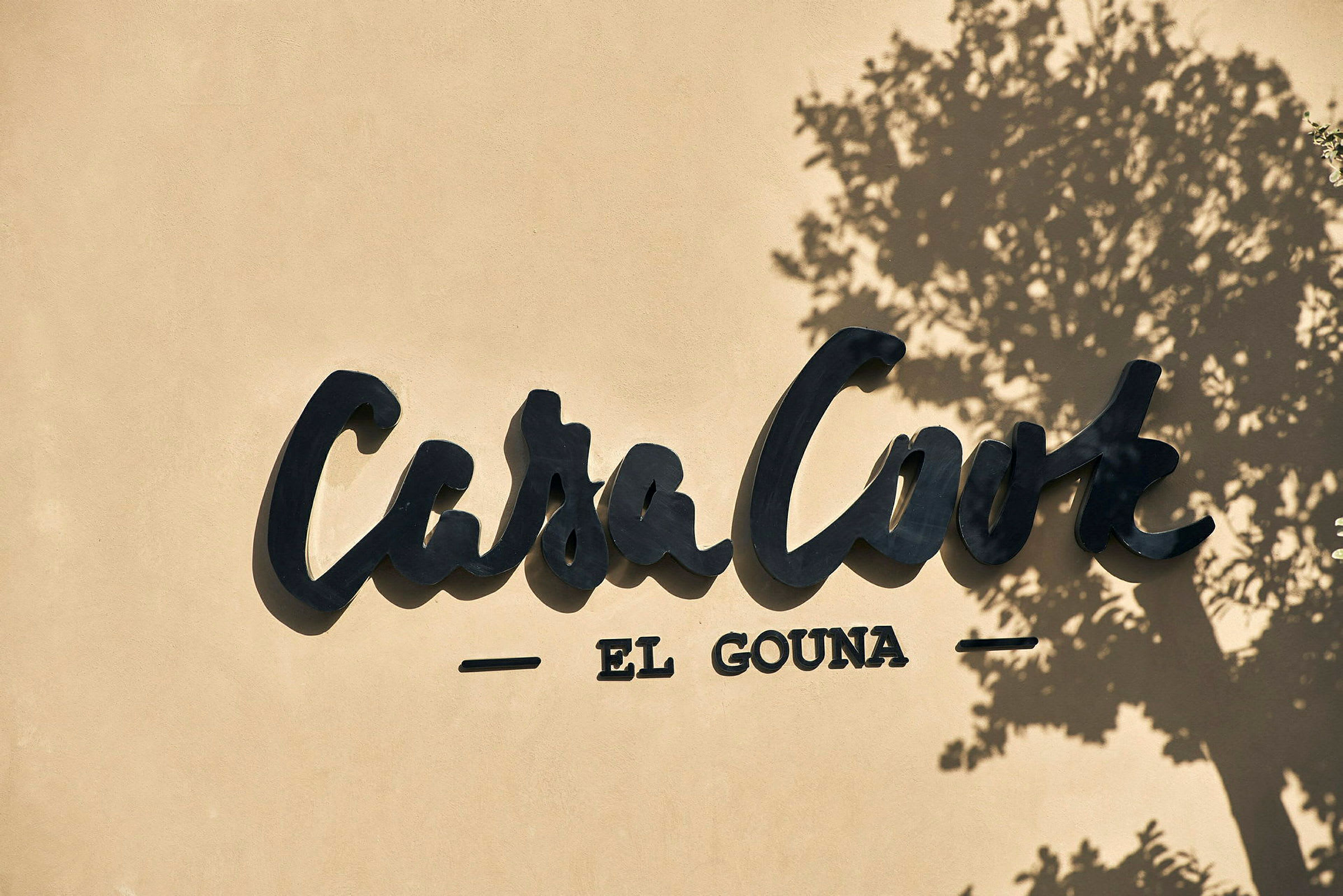 Logotypen för Casa Cook El Gouna på en vägg, med skuggan av ett träd. (AI-genererad text)