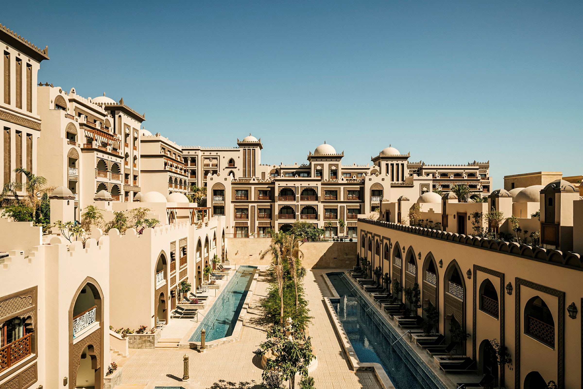 Bilden visar ett hotellkomplex med arkitektur inspirerad av arabisk stil, inklusive domer och ornamenterade valv, samt två långsmala pooler omgivna av byggnader. (AI-genererad text)