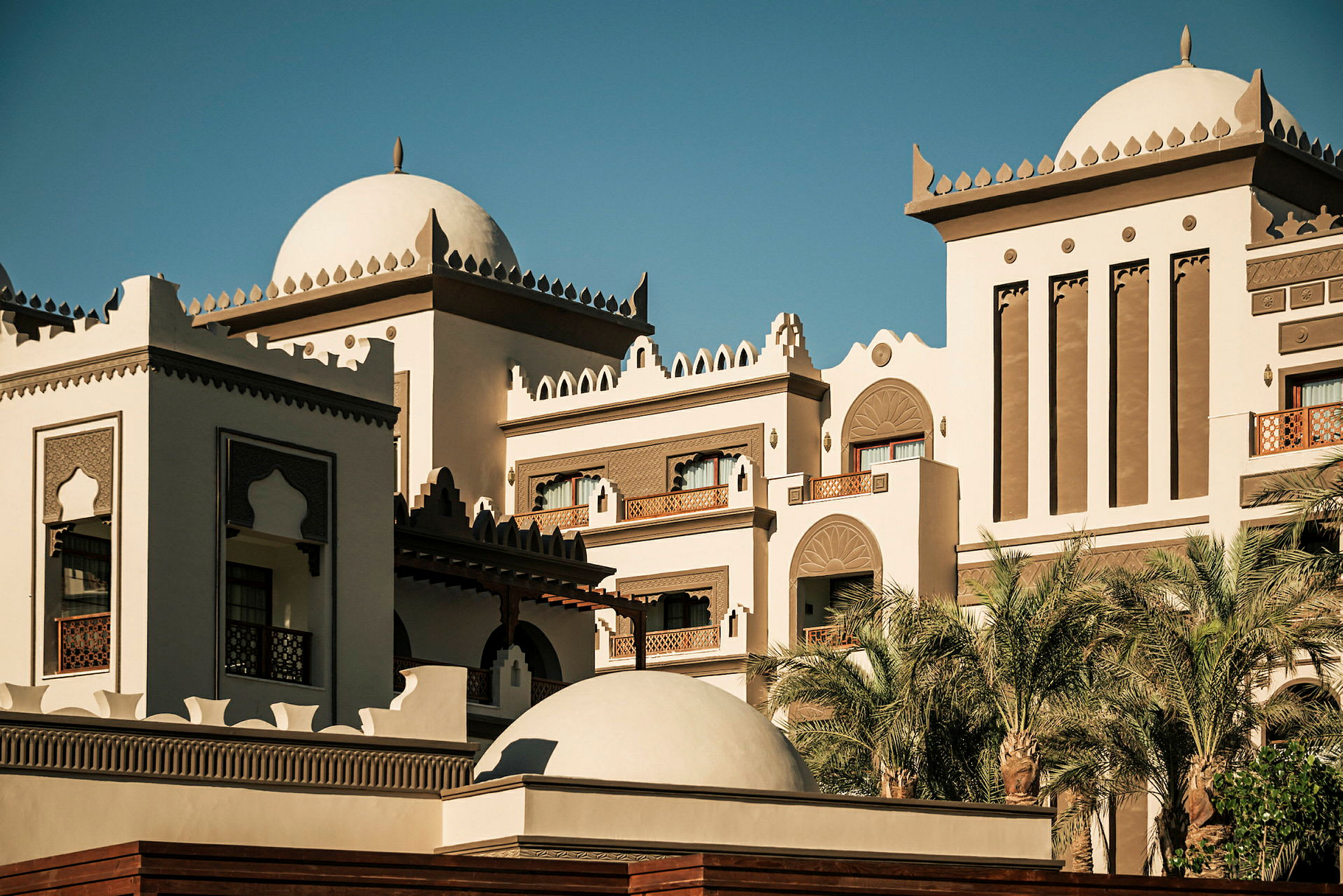 En byggnad i orientaliskt arkitektonisk stil med kupoltak och dekorativa detaljer, omgiven av palmer och blå himmel. (AI-genererad text)