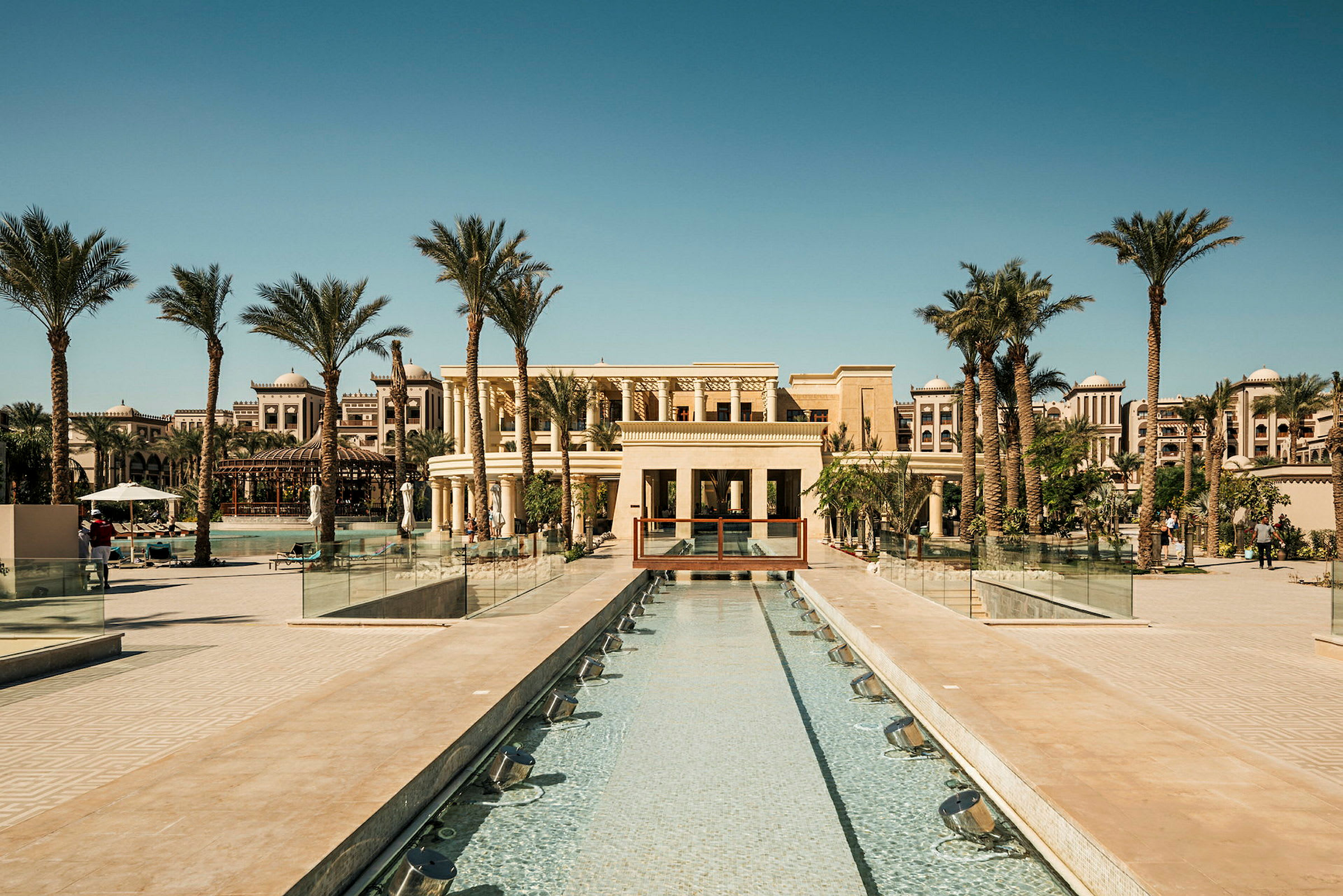 Bild av en lyxig resort med en lång grund pool omgivet av höga palmträd, moderna byggnader och sittmöbler. (AI-genererad text)