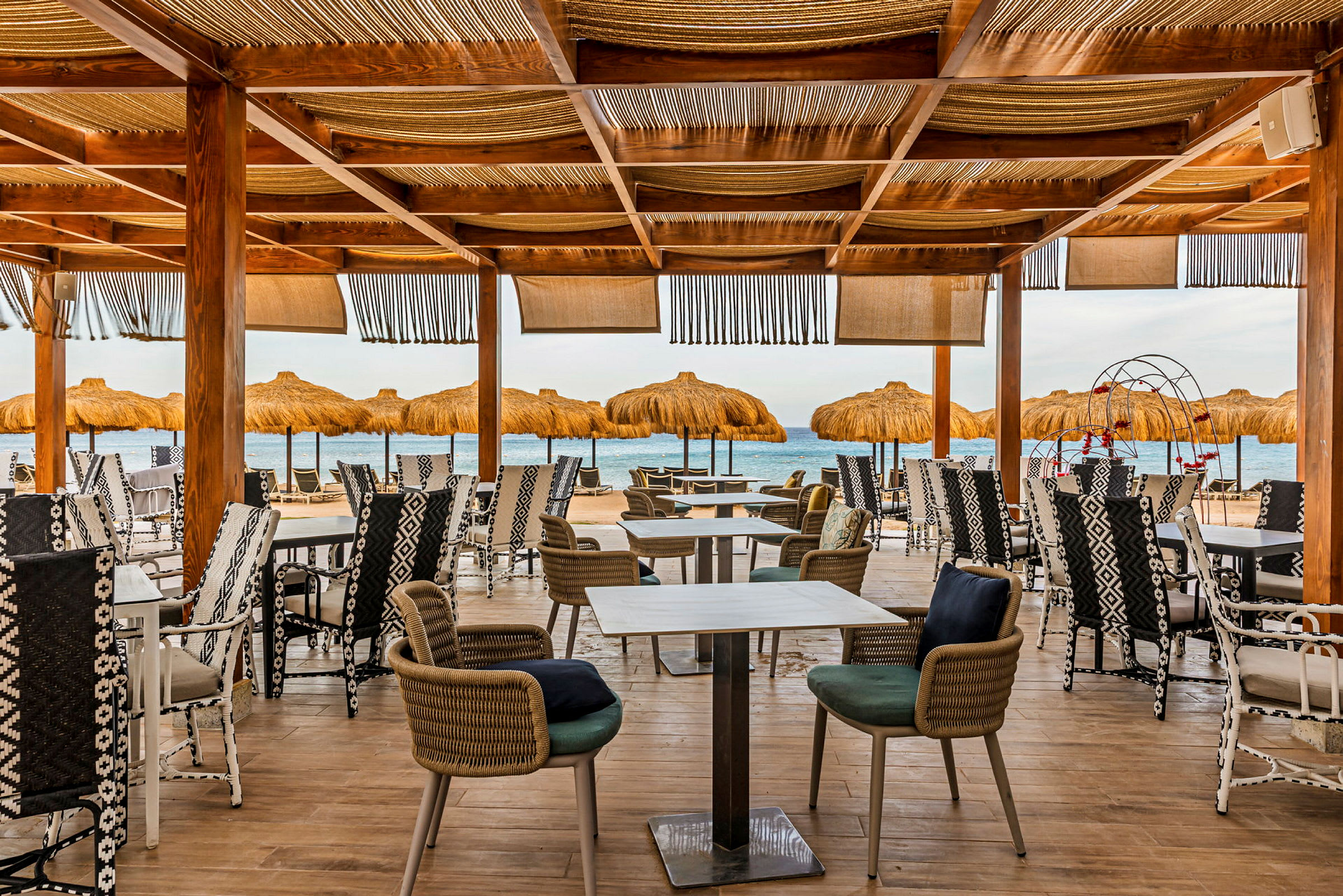 En utomhusrestaurang med träpelare och stolar, med utsikt över havet och parasoller på stranden. (AI-genererad text)