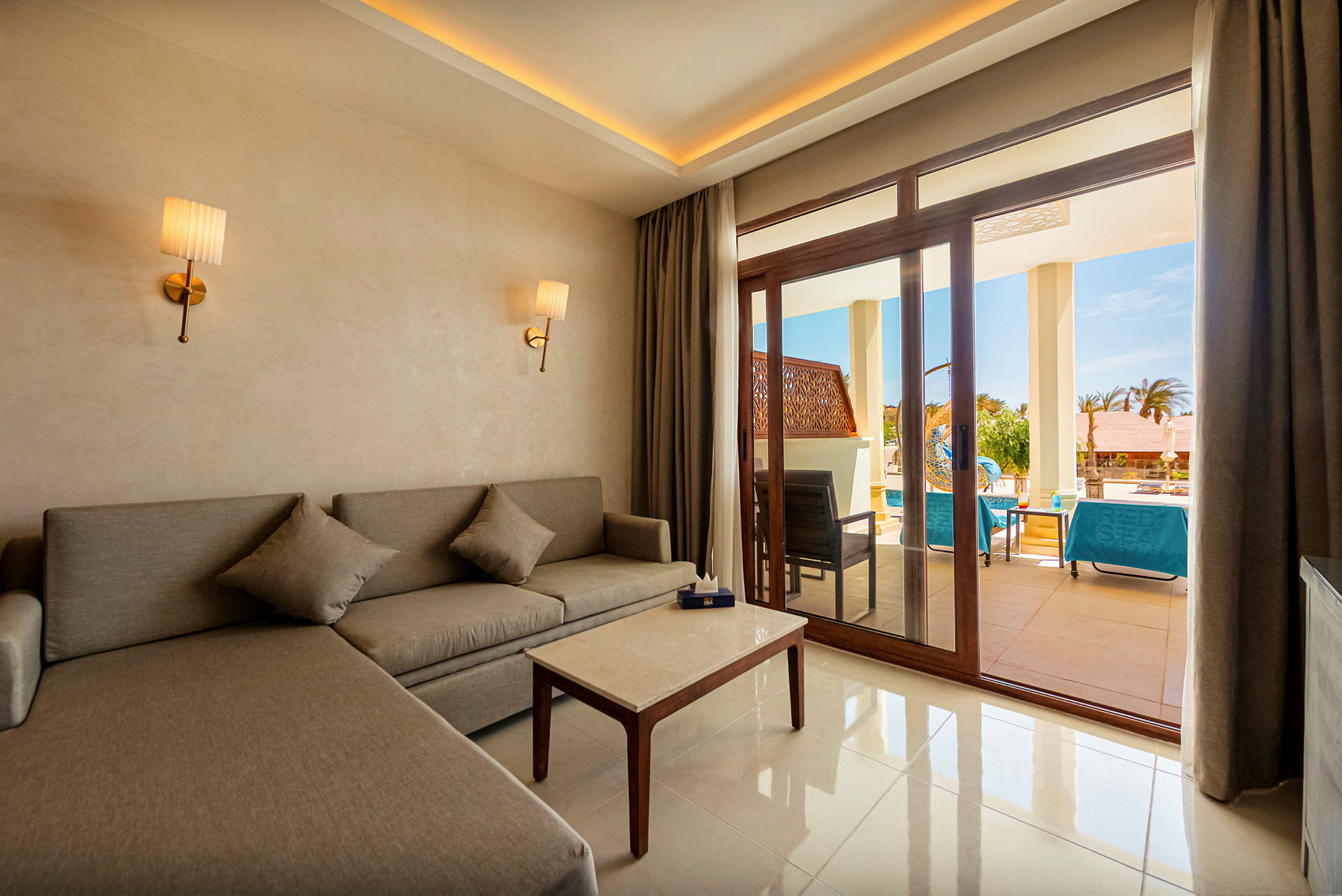 En hotellsvit med en grå soffa, ett soffbord och två vägglampor. Utanför glasdörrarna syns en terrass med sittmöbler och solsängar vid en pool. (AI-genererad text)