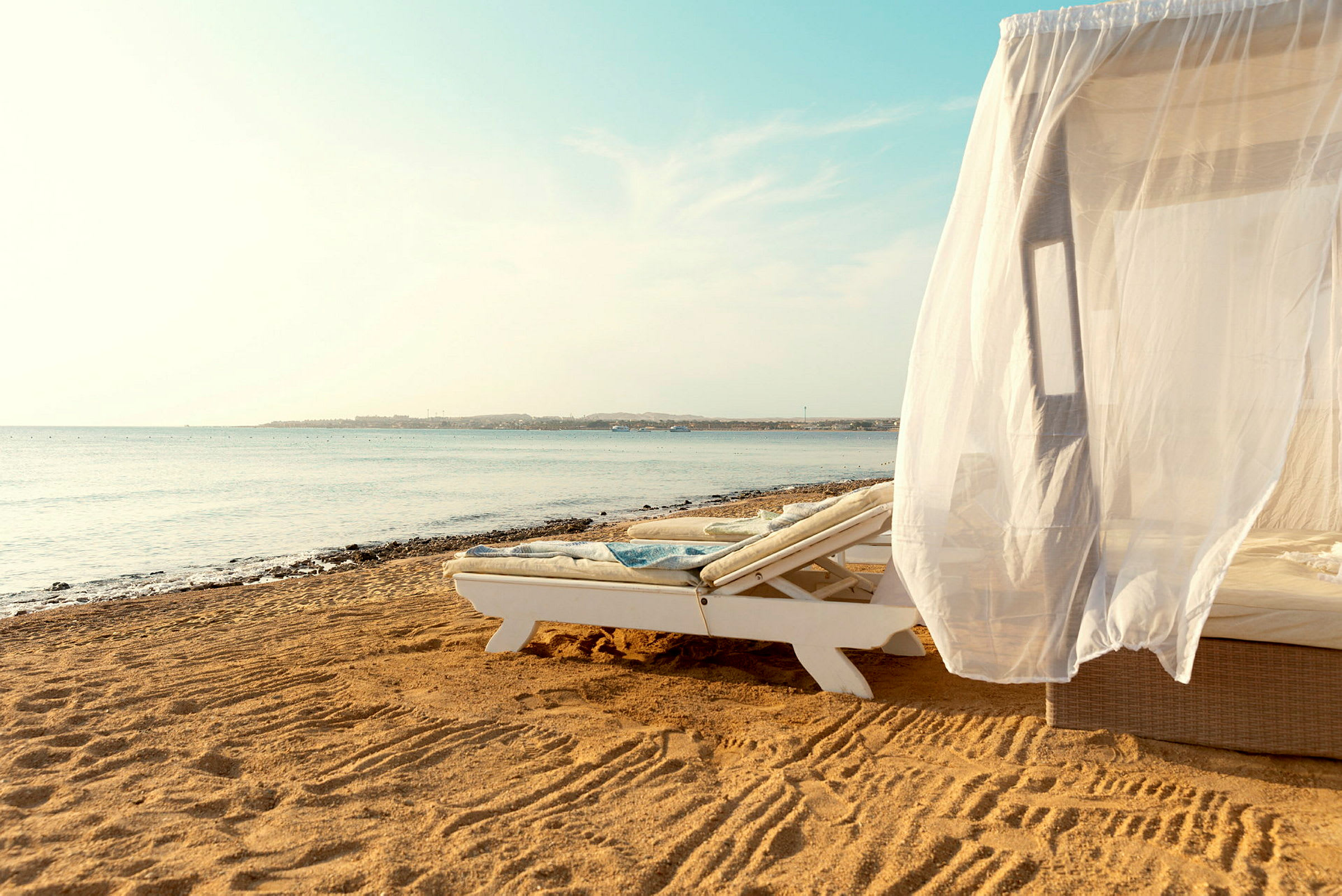 En solsäng med ett vitt draperi på en sandstrand vid havet. (AI-genererad text)