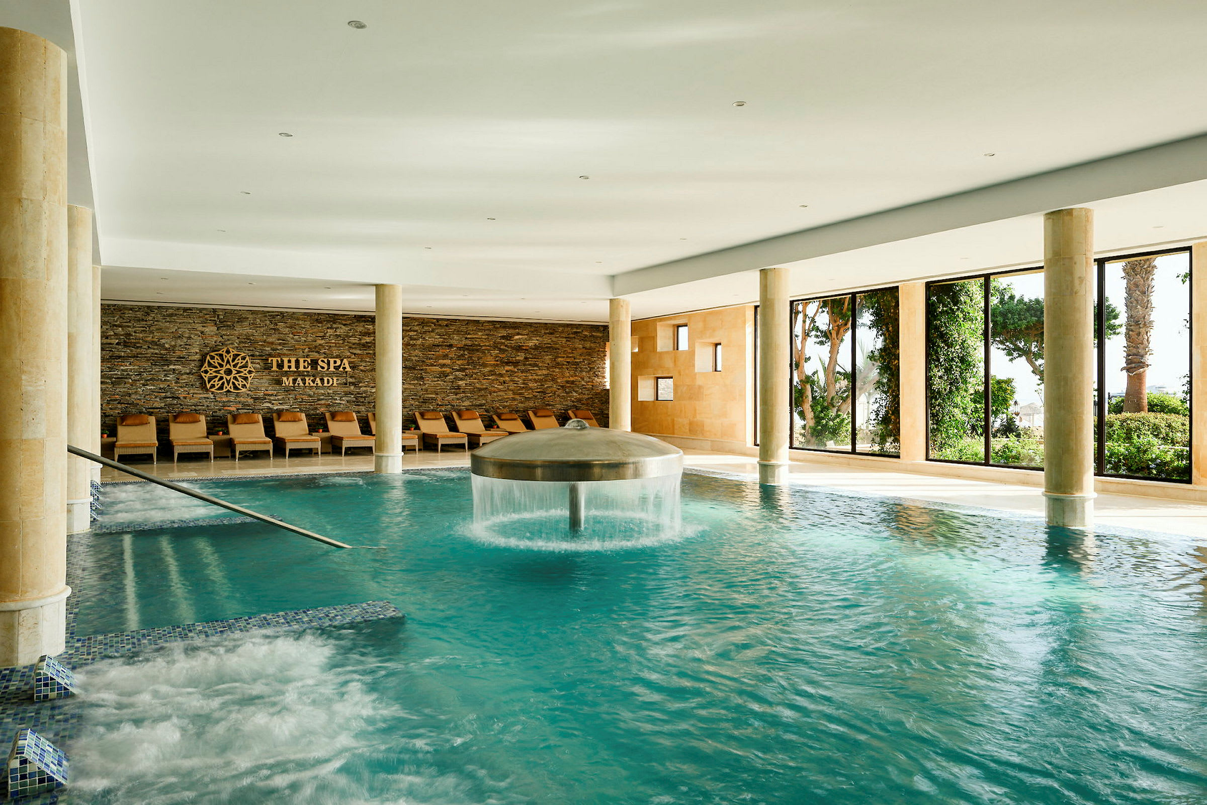 Ett lyxigt spa med en inomhuspool. Poolområdet har stora fönster som släpper in naturligt ljus, och på ena sidan finns ett antal vilstolar vid en vägg med dekorativ stenbeklädnad. (AI-genererad text)