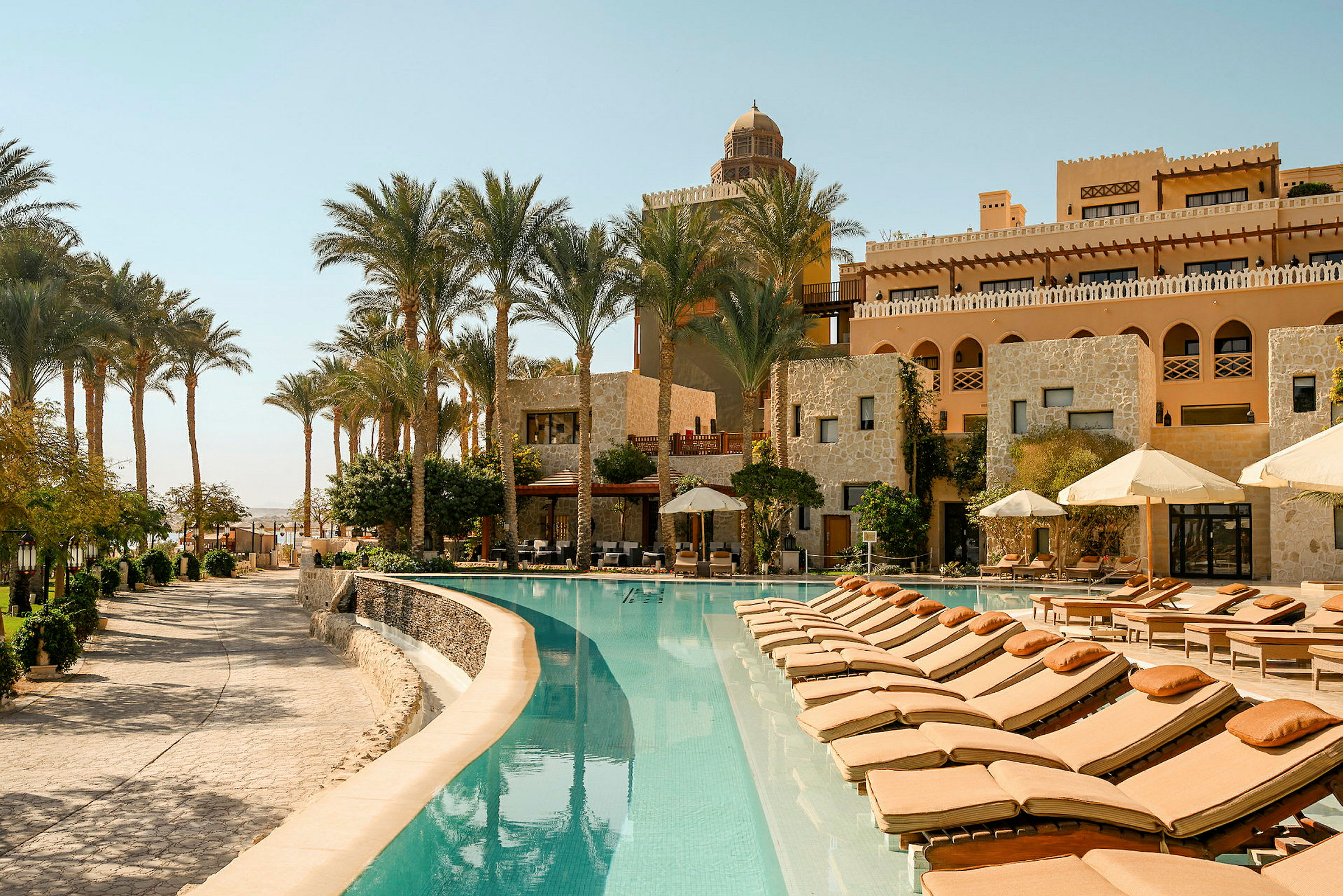 Ett elegant hotellområde med en pool omgiven av solstolar och parasoller. Byggnader i bakgrunden är byggda i sten och dekorerade med bågformade fönster. Palmträd omger området. (AI-genererad text)
