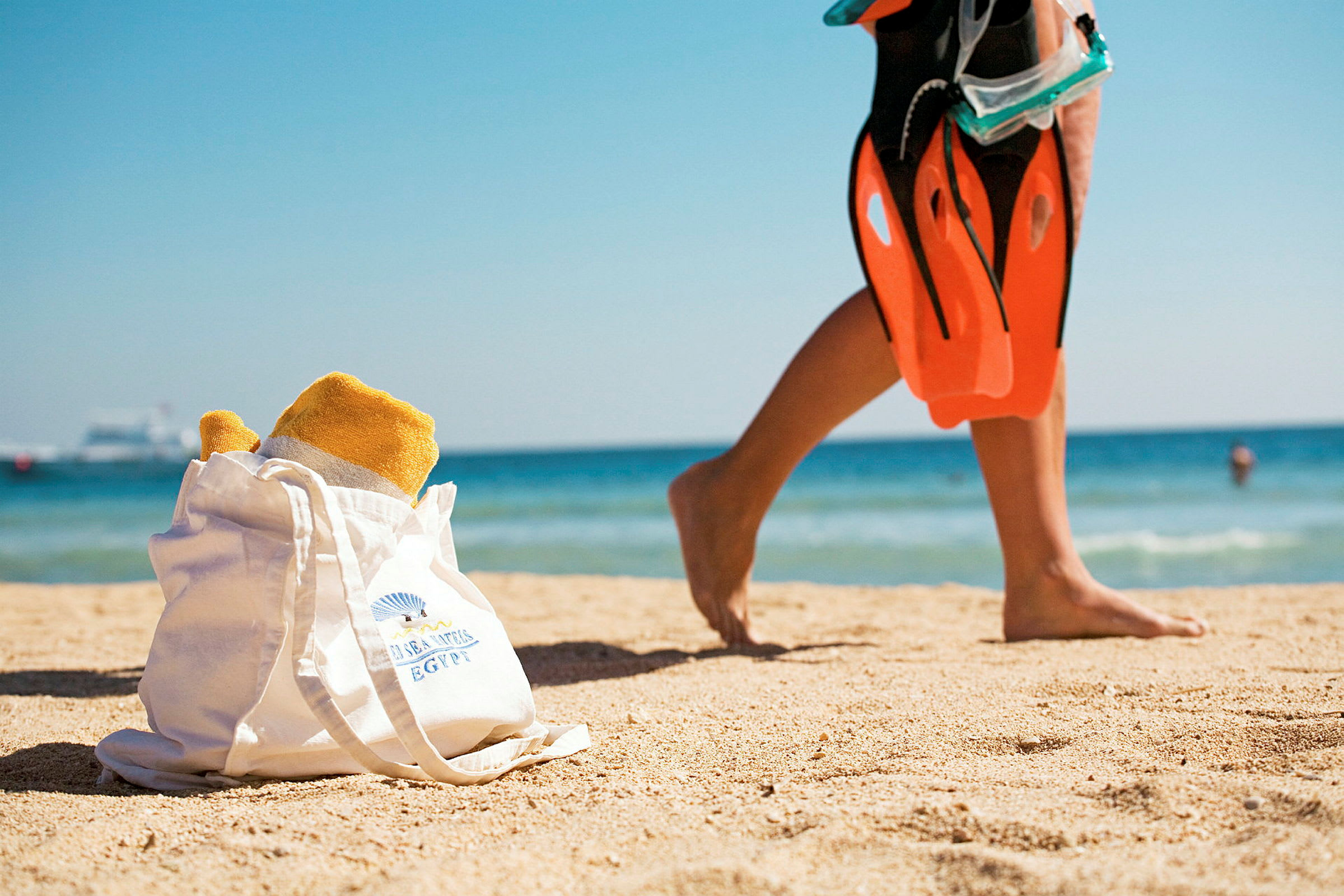 Strand med vit väska innehållande handduk i förgrunden och person med simfenor i bakgrunden. (AI-genererad text)
