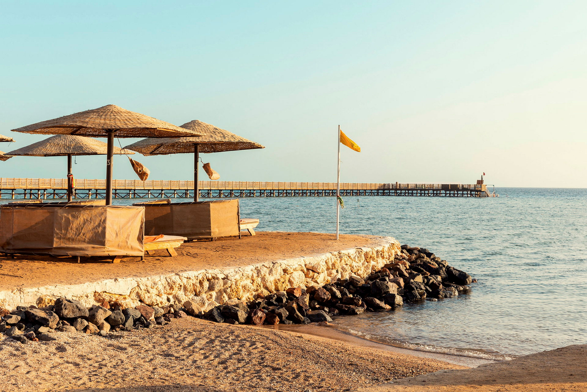 Strand med parasoller och solstolar, samt en brygga som sträcker sig ut i havet. (AI-genererad text)