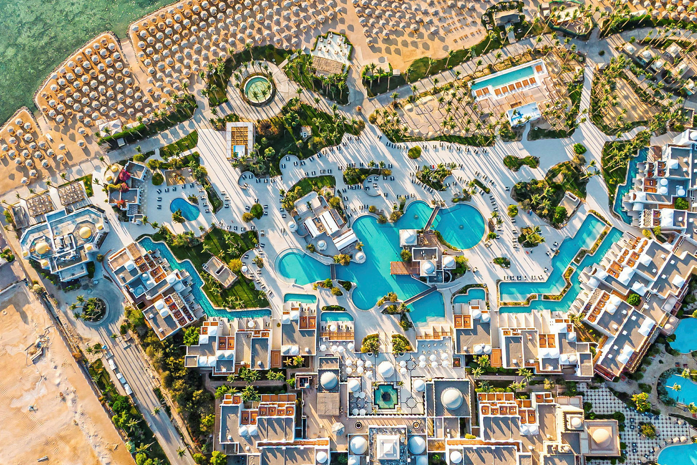 Flygvy över ett semesterkomplex med pooler, byggnader och en strand med parasoll. (AI-genererad text)