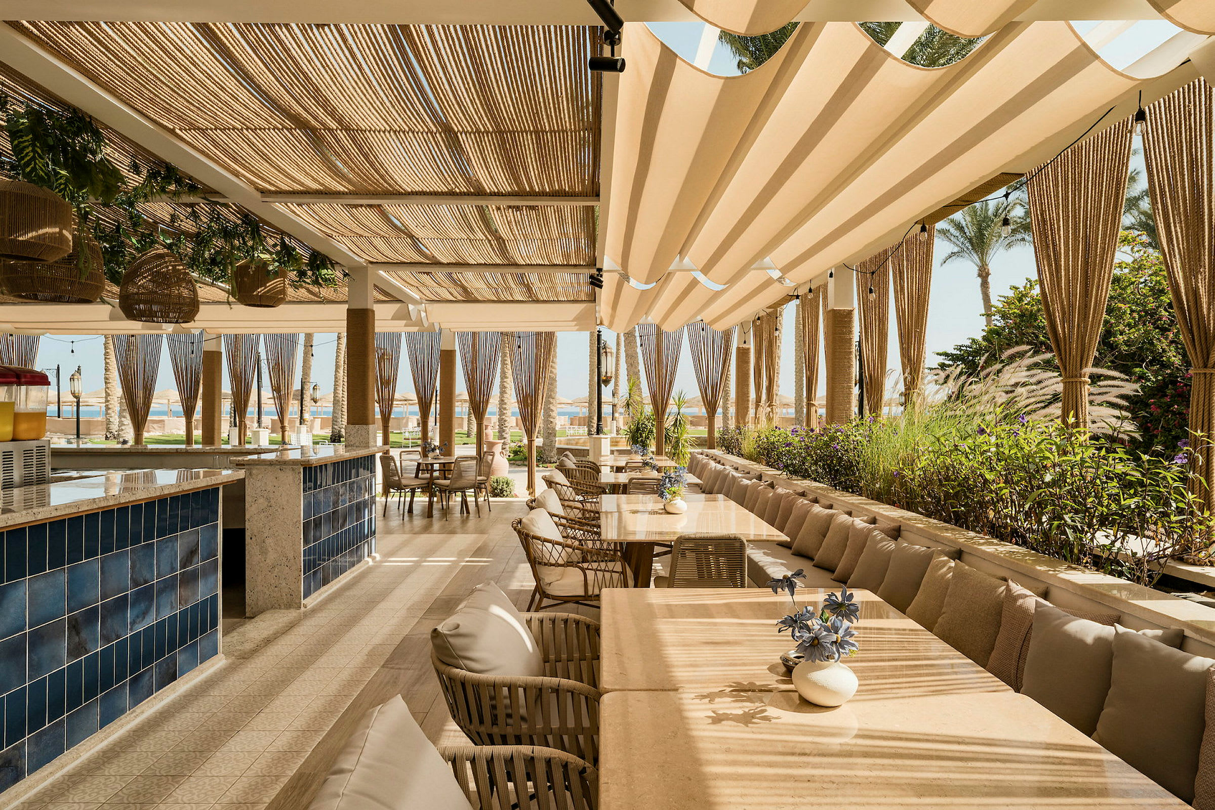 En restaurangterrass med bord, stolar och beige solskydd, omgiven av gröna växter. (AI-genererad text)