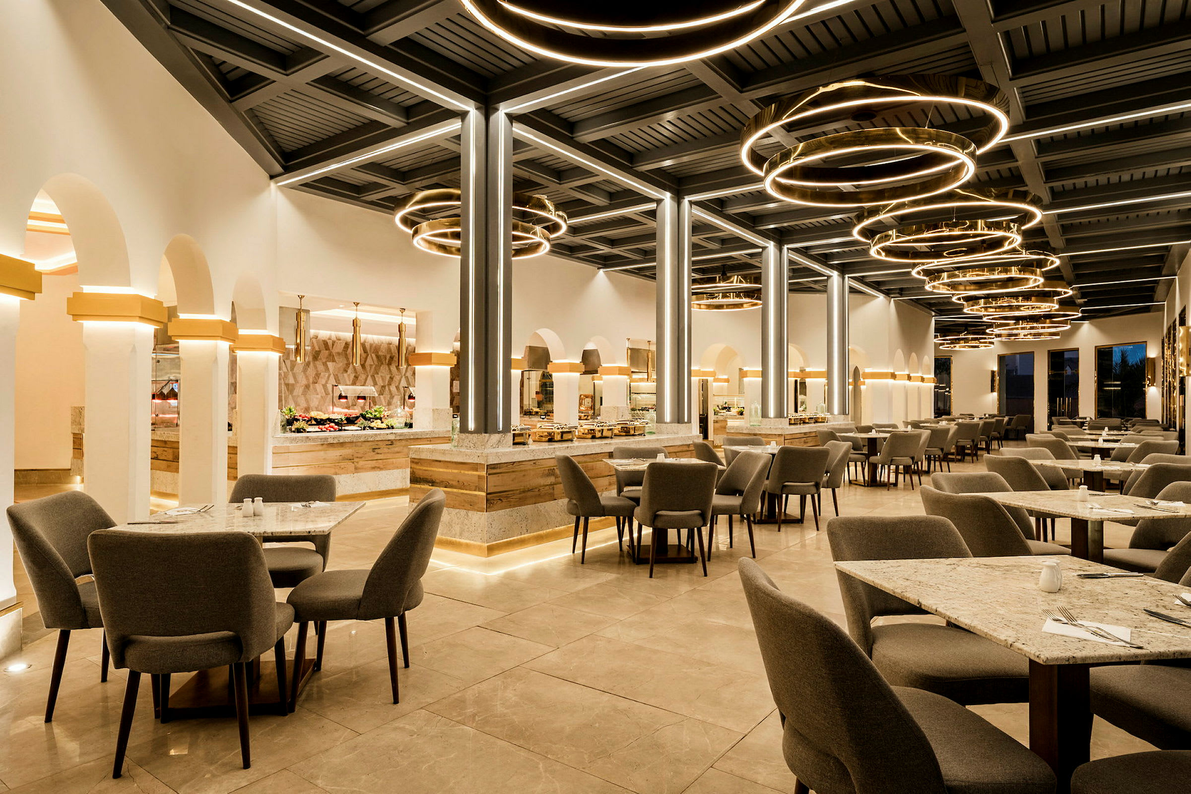 En modern restaurang med grå stolar och bord på ett stenlagt golv, lysande cirkelformade lampor hänger från taket. (AI-genererad text)
