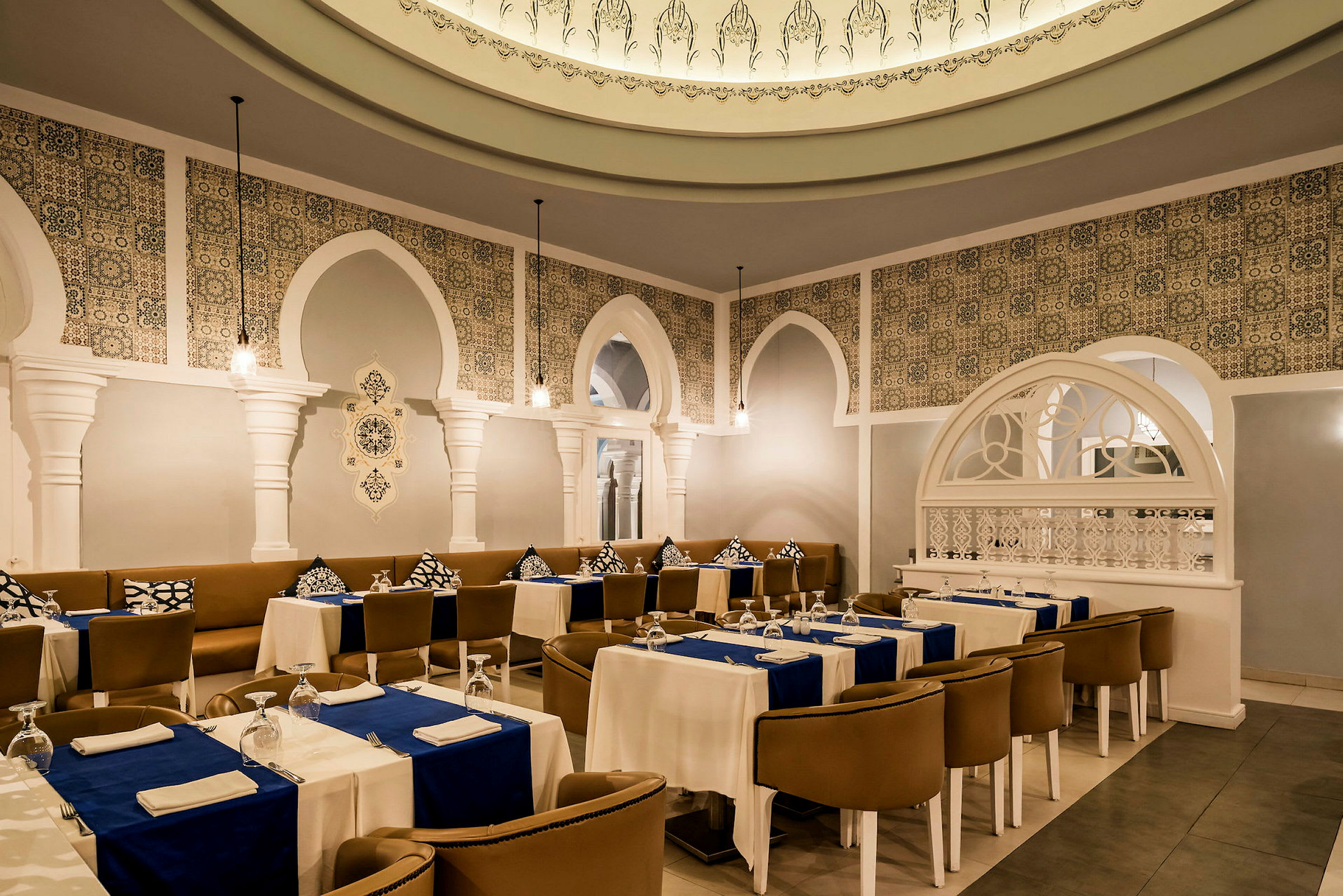 En elegant restaurang med vita kolonner, dekorativ kakelmönster och uppdukade bord med blå bordsdukar. (AI-genererad text)