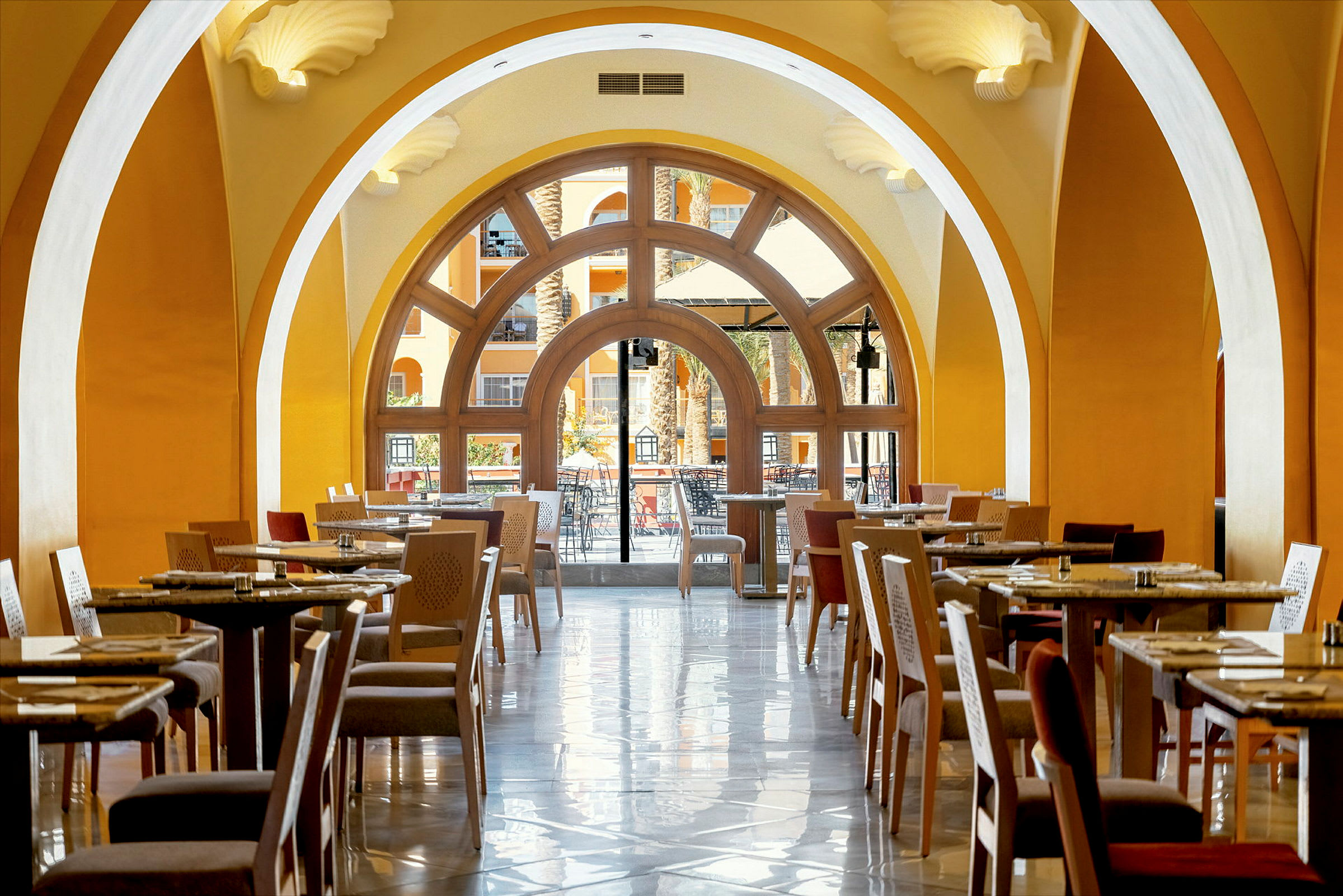 En restaurang med sittplatser och bord under välvna tak med utsikt mot en terrass genom stora fönster. (AI-genererad text)