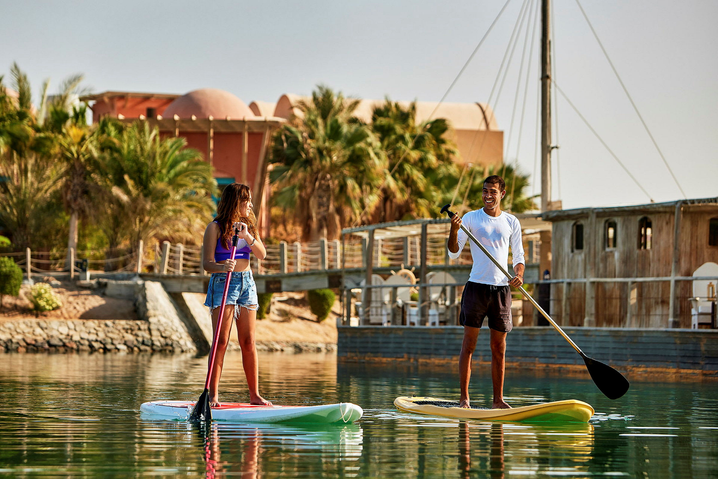 Två personer står på paddleboards vid vattnet med palmer och byggnader i bakgrunden. (AI-genererad text)