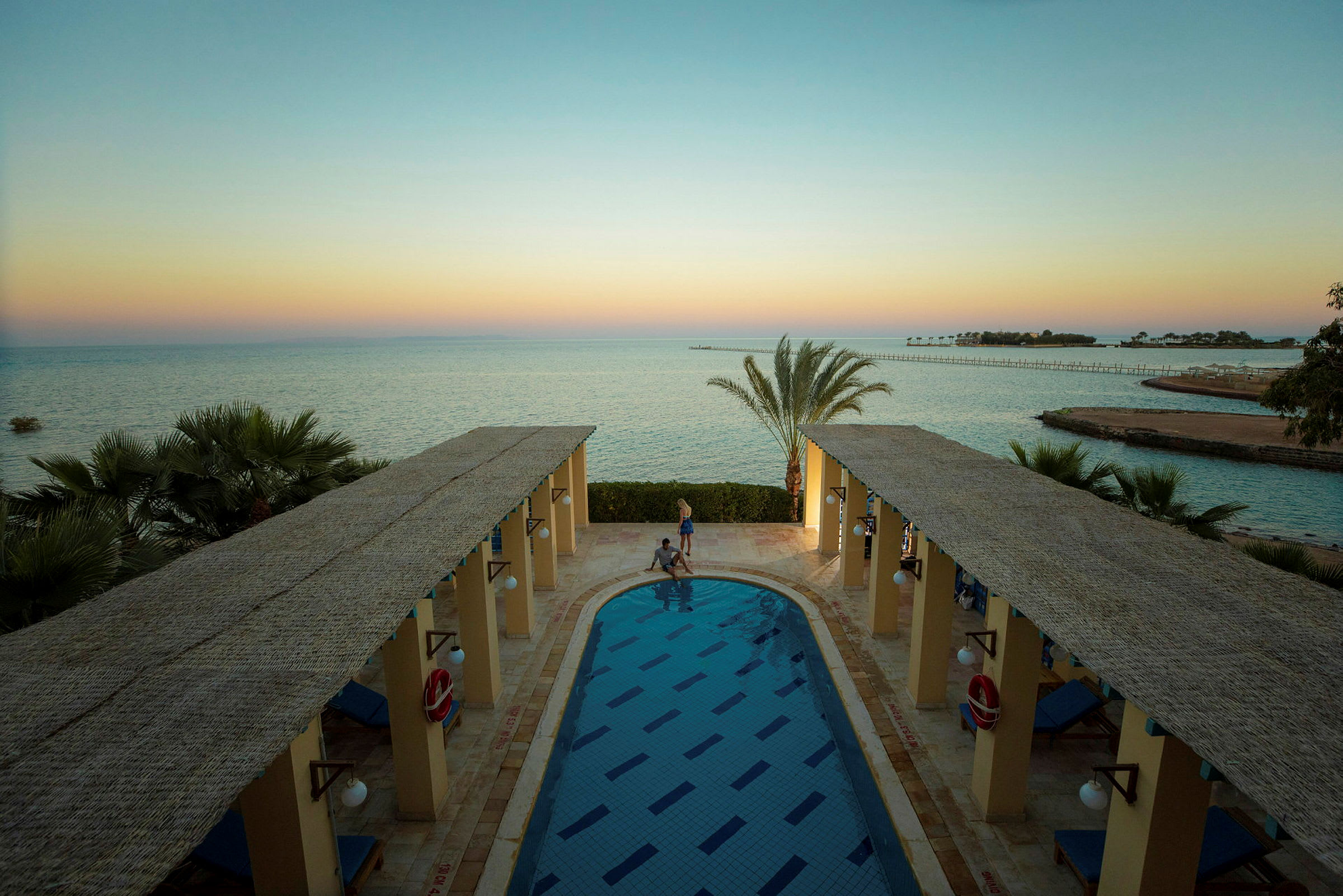 Utsikt över en lång pool mellan två terrasser med tak, med havet och en brygga i bakgrunden. (AI-genererad text)