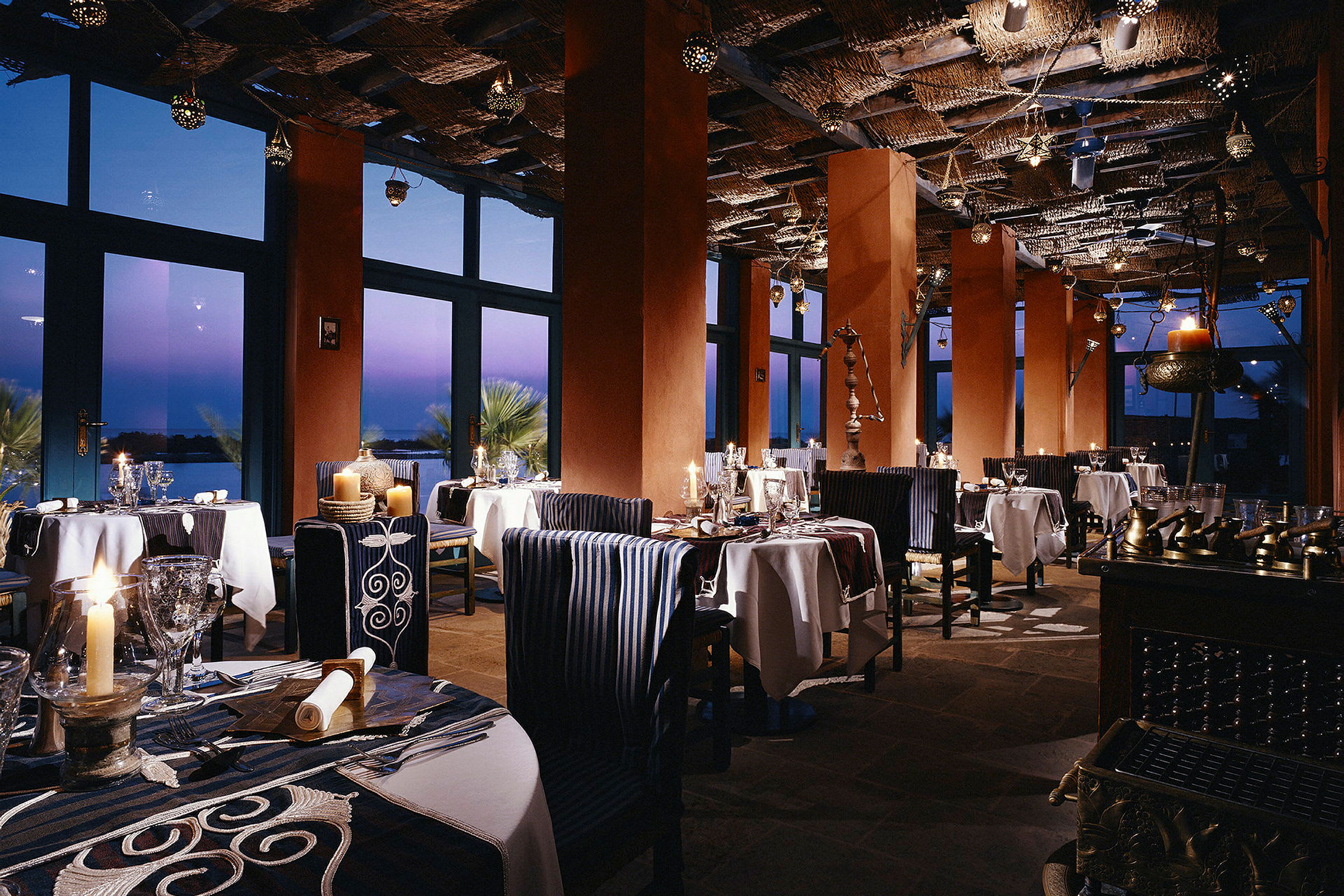 Restaurang med belysning, bord och stolar, samt stora fönster mot skymningen. (AI-genererad text)