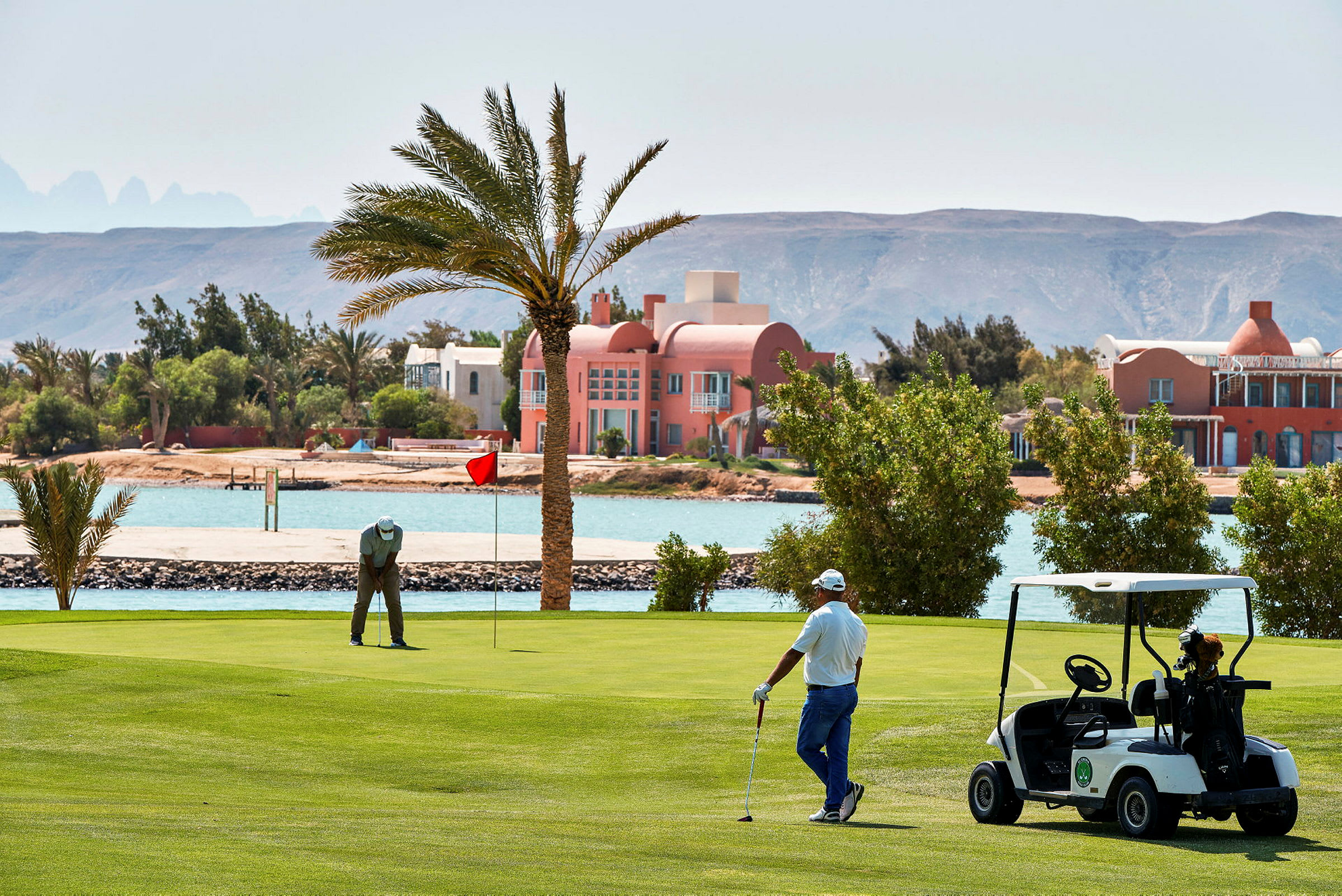 Två personer spelar golf på en grön golfbana nära vattnet, med ett palatsliknande hus i bakgrunden. (AI-genererad text)