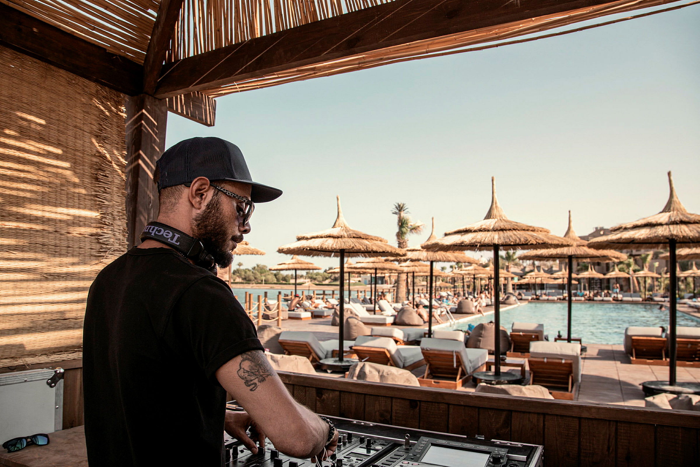 En DJ vid sitt mixerbord med en bakgrund av solstolar och parasoller vid en pool. (AI-genererad text)