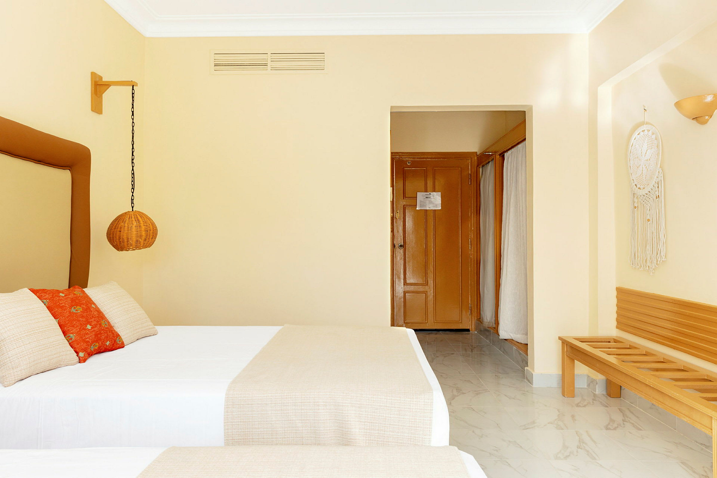 Ett hotellrum med två enkelsängar, dekorativ korgpendel, träbänk, och en drömfångare på väggen. Dörren till badrummet syns i bakgrunden. (AI-genererad text)