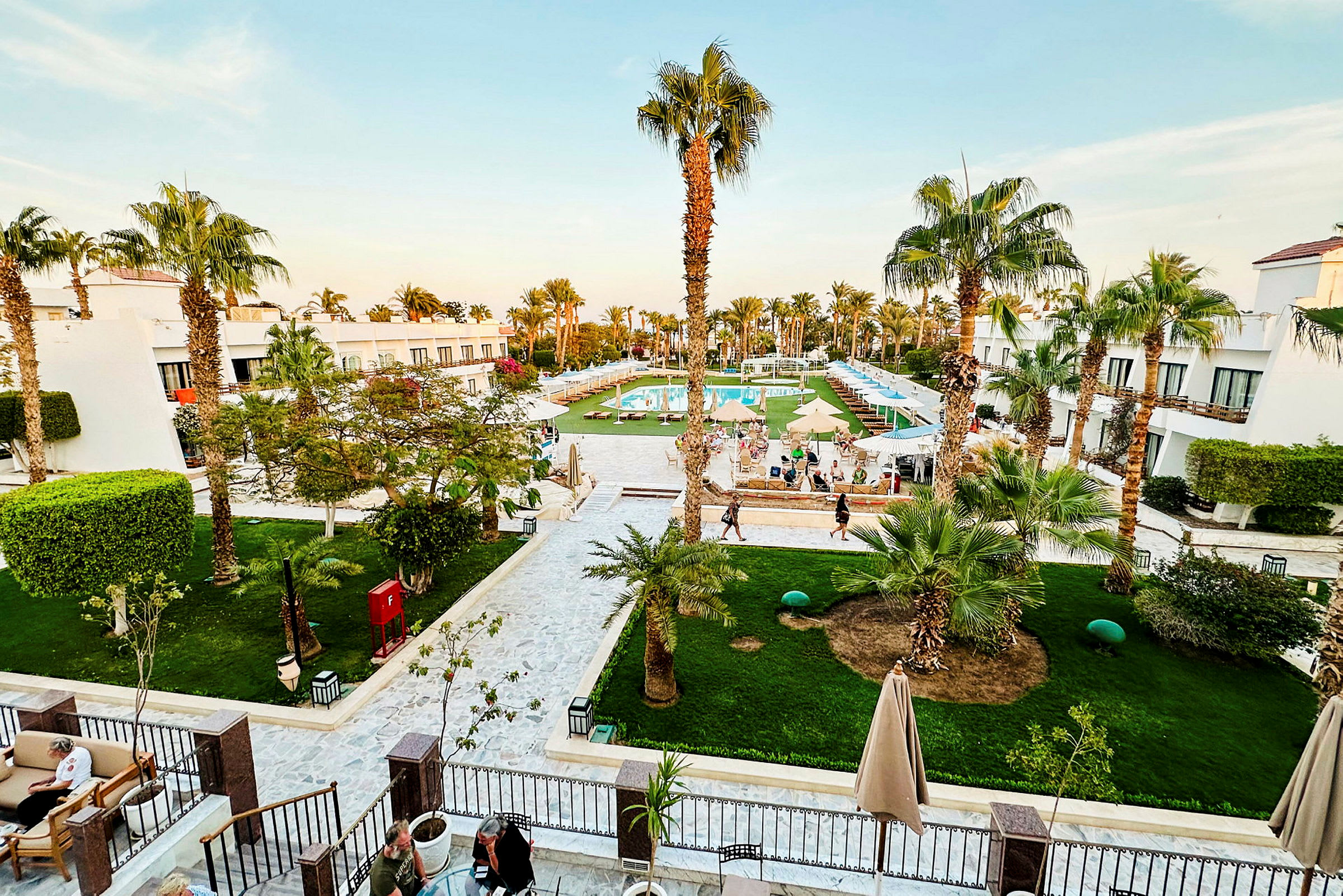 Vy över ett hotellområde med gröna gräsmattor, palmer, vita byggnader och en pool i mitten. (AI-genererad text)