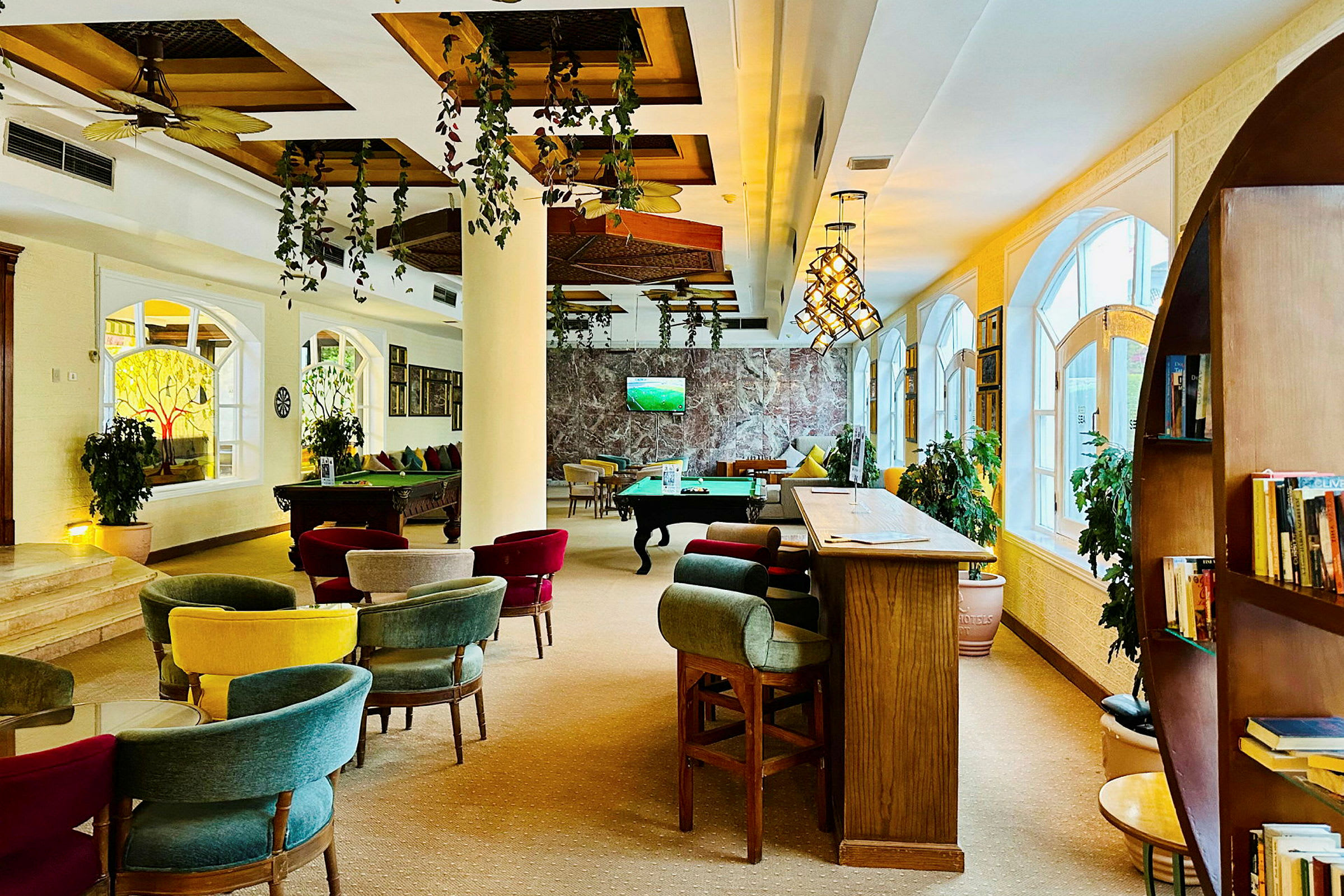 Ett inomhusutrymme med sittgrupper, biljardbord och en bar med höga sittplatser. Växter och stora fönster dekorerar rummet. (AI-genererad text)